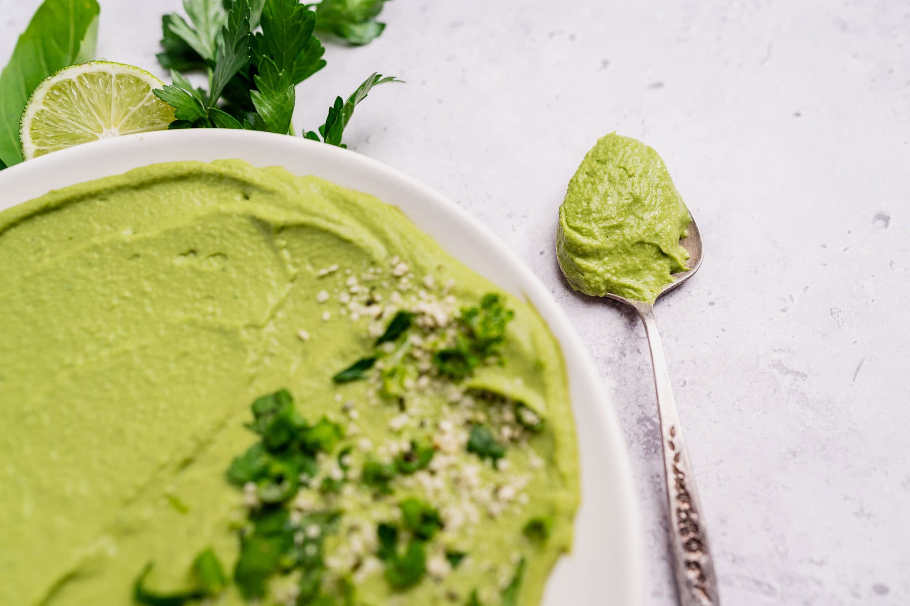 A plate of green goddess hummus topped with herbs and seeds sits next to a spoonful of hummus, with fresh greens and a lime wedge in the background.