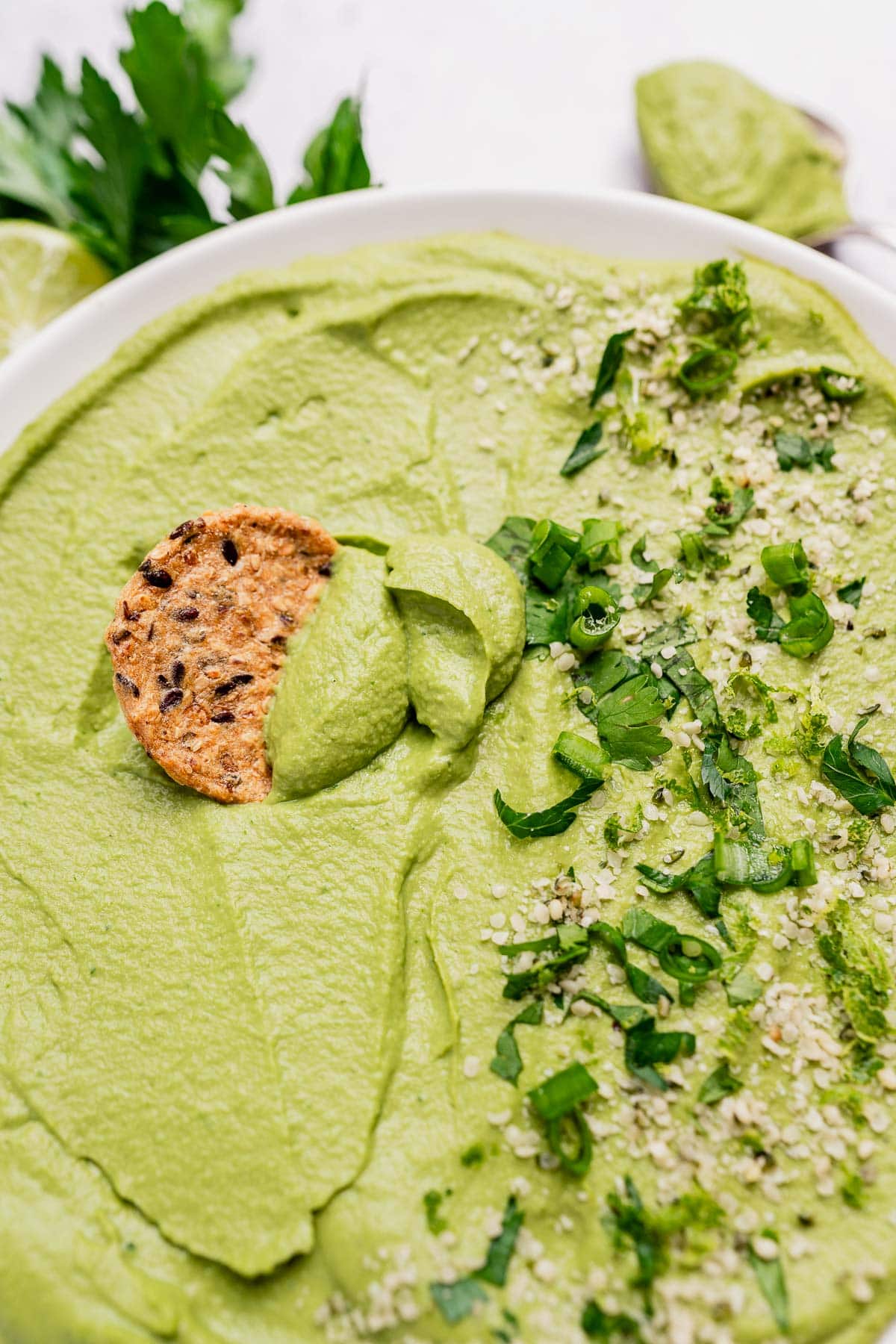 A bowl of creamy green goddess hummus garnished with chopped herbs and seeds, with a seeded cracker partially dipped into it.