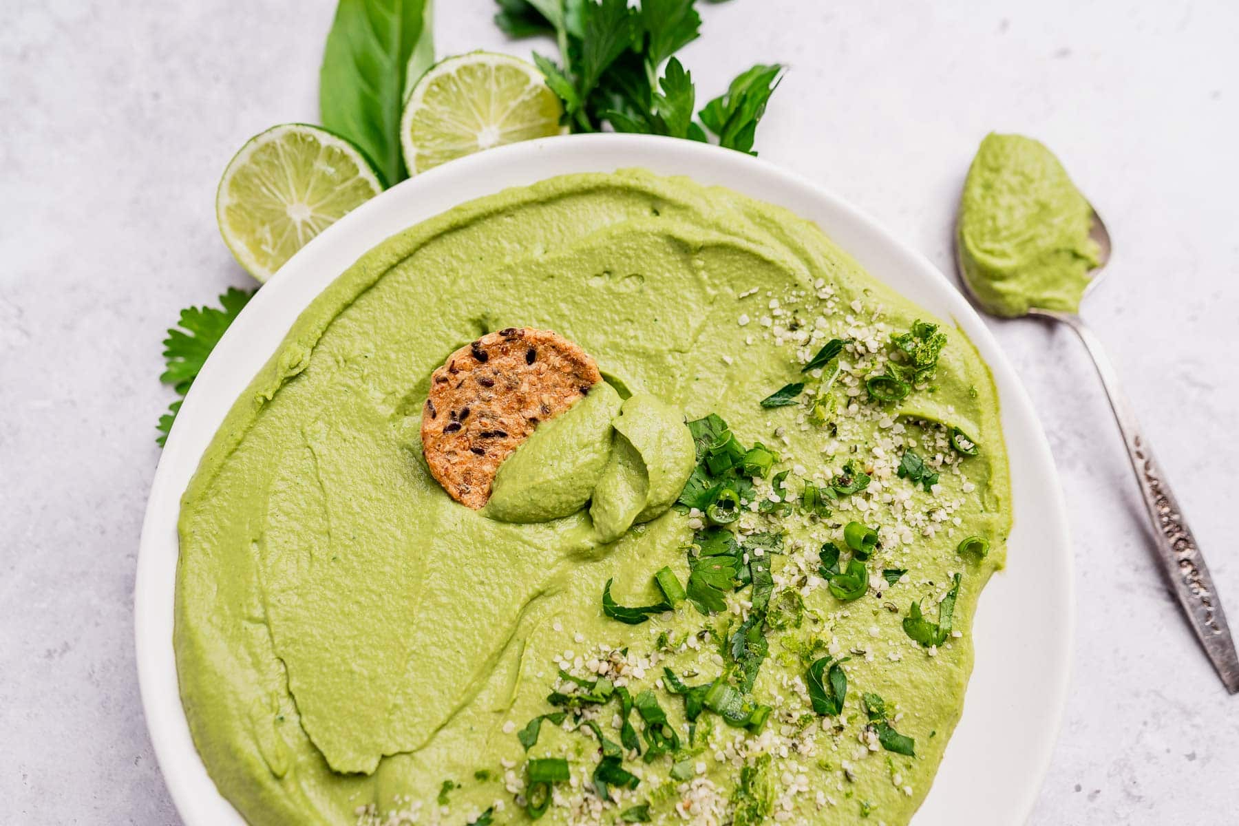 A white bowl filled with vibrant green goddess hummus, topped with chopped herbs and a cracker, with lime slices and fresh herbs nearby and a spoonful of hummus on the side.