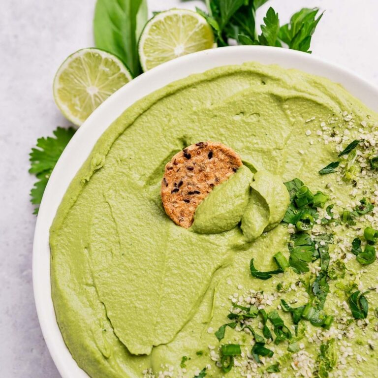 A bowl of green goddess hummus garnished with chopped herbs and seeds, with a round cracker partially dipped in the creamy dip. Slices of lime and fresh herbs add brightness in the background.