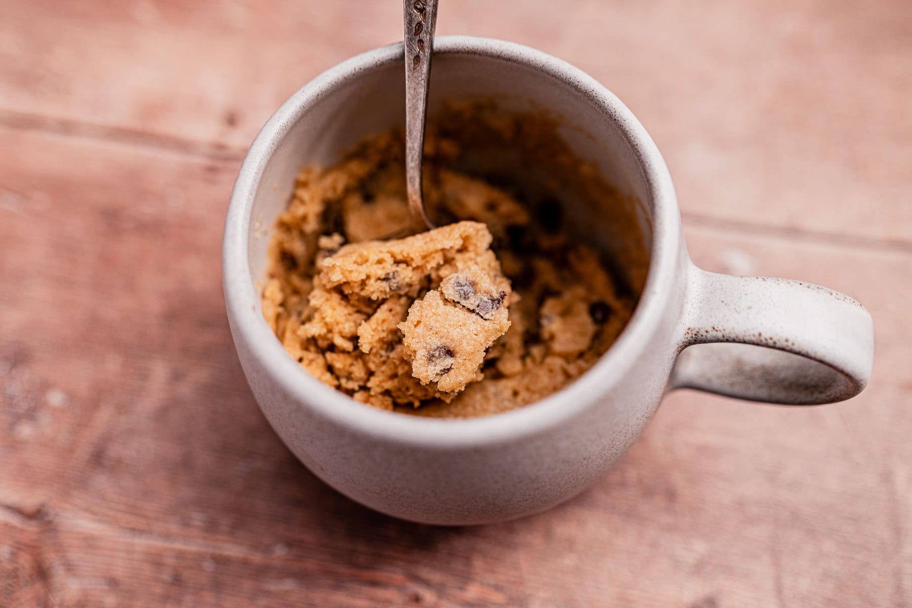 A mug filled with chocolate chip cookie dough and a spoon rests on a wooden surface, hinting at the perfect oat flour mug cake just waiting to be devoured.