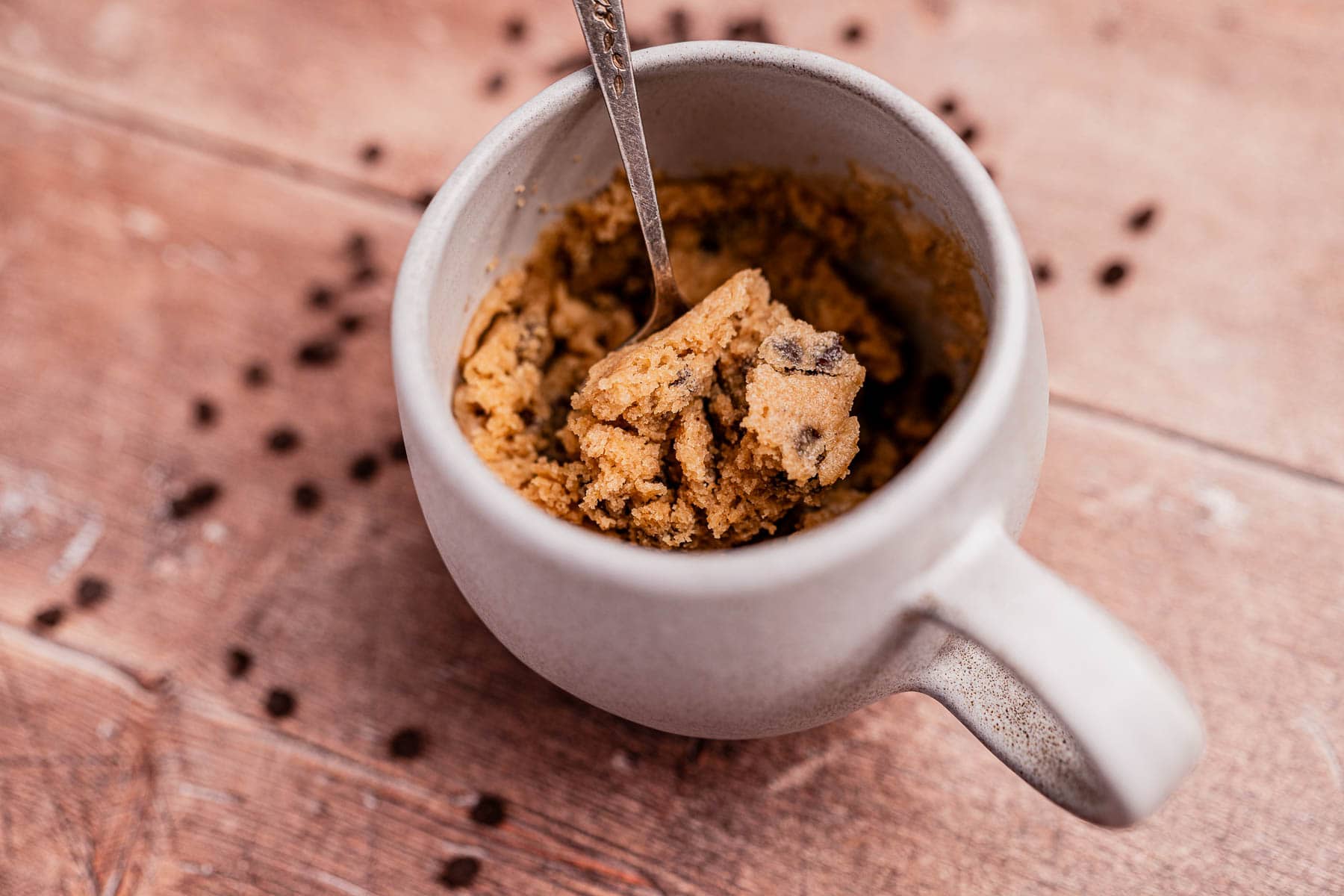 A ceramic mug filled with oat flour mug cake cookie dough and a spoon, with chocolate chips scattered on a wooden surface around the mug.