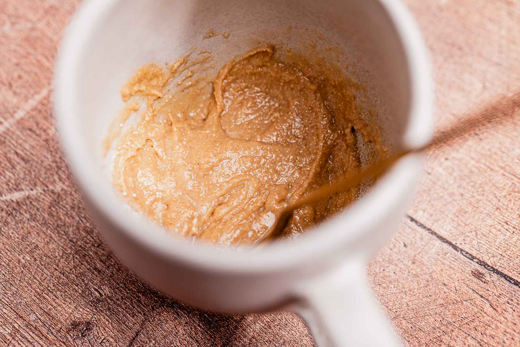 A white mug containing a thick, brown oat flour batter with a spoon inside, placed on a wooden surface.