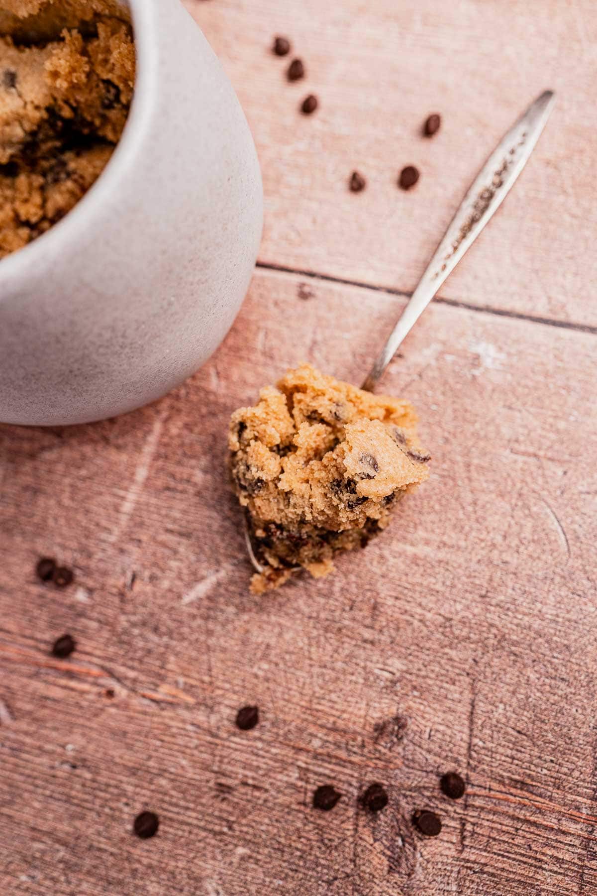 A spoonful of chocolate chip cookie dough made with oat flour mug cake mix rests on a wooden surface, with a jar of more dough and scattered chocolate chips nearby.