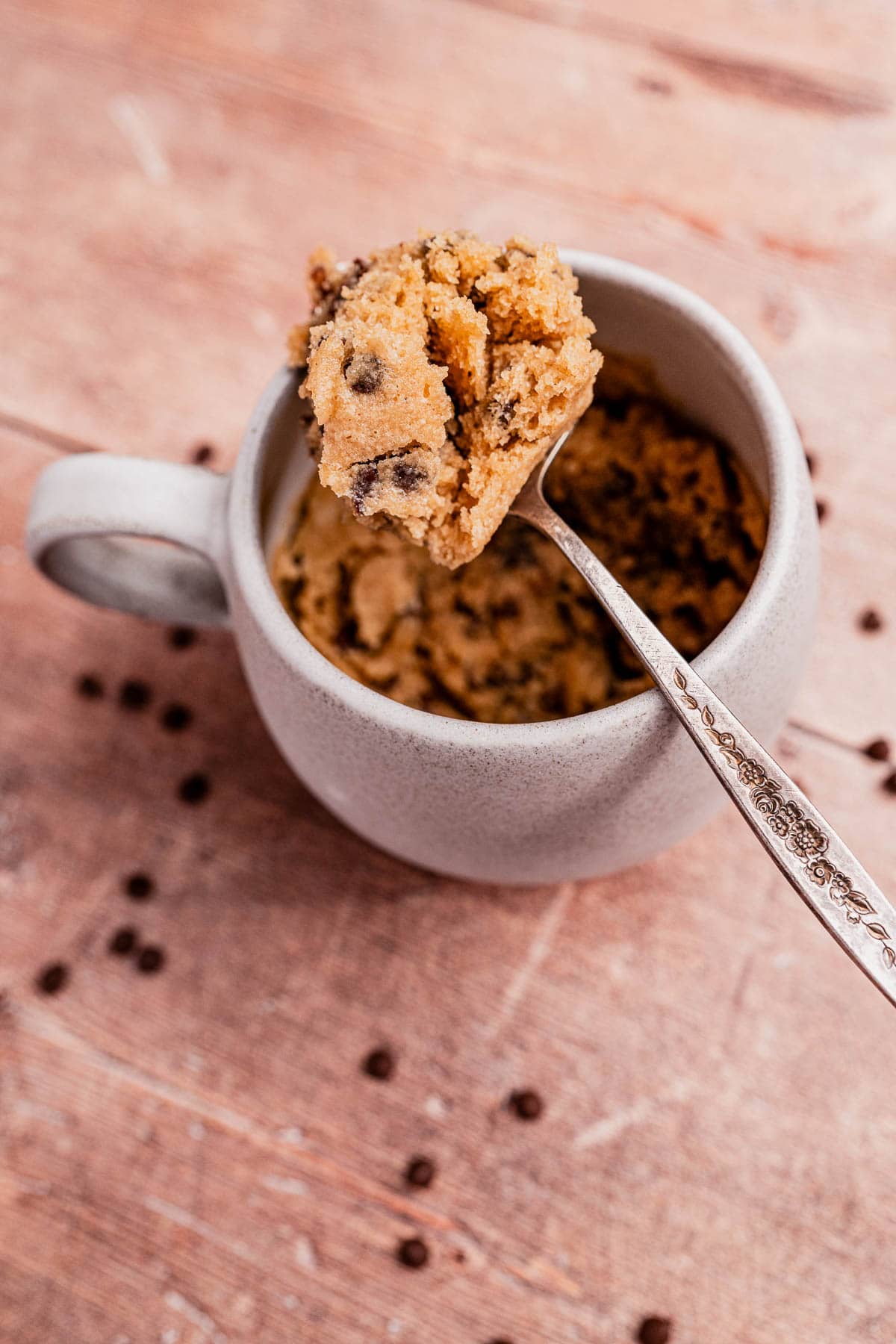 A spoon holds a portion of chocolate chip oat flour mug cake dough above a mug filled with more dough, with scattered chocolate chips on a wooden surface.