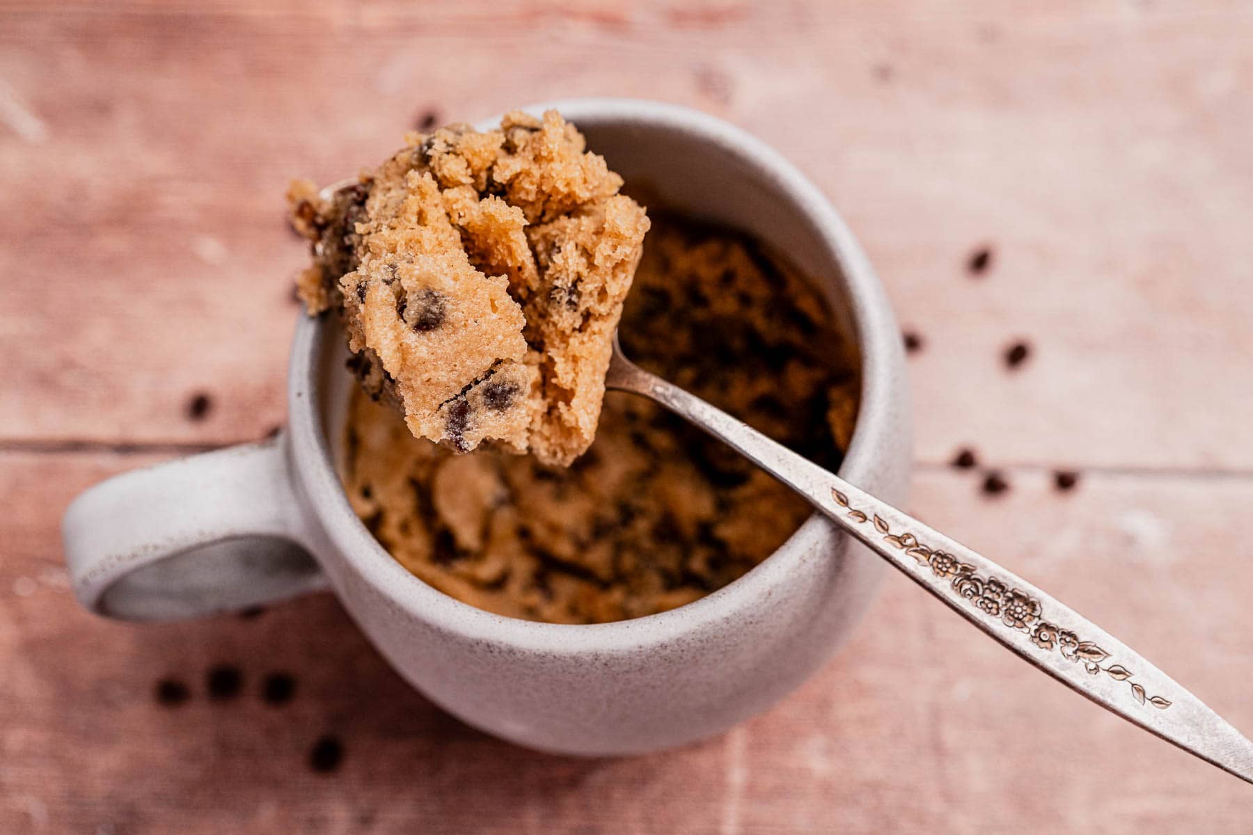 A spoon holds a portion of oat flour mug cake with chocolate chips above a white ceramic mug on a wooden surface.
