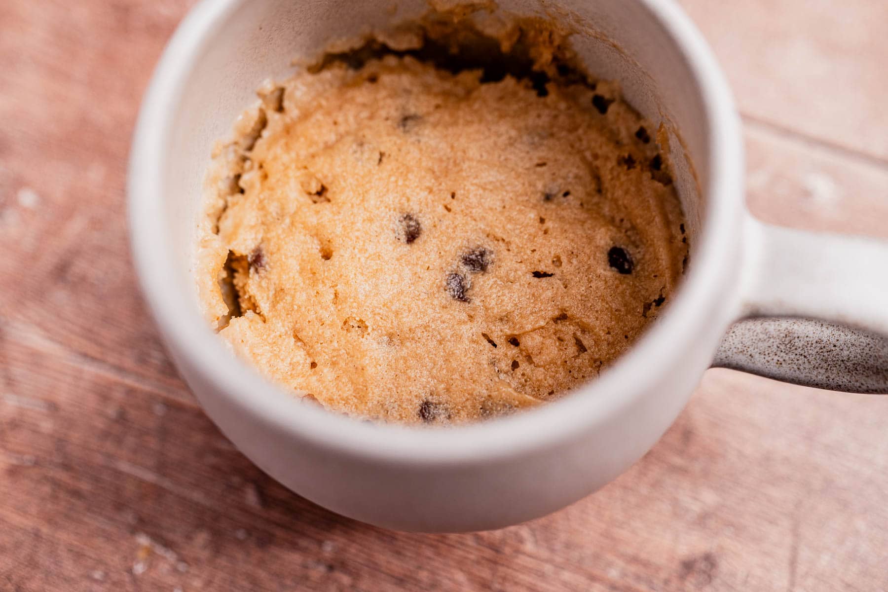 A delightful oat flour mug cake with chocolate chip cookie dough, nestled in a beige mug on a wooden surface.