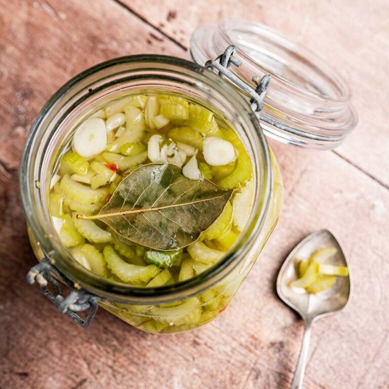 An open glass jar of pickled vegetables, including pickled celery and topped with a bay leaf, sits next to a spoon on a wooden surface.