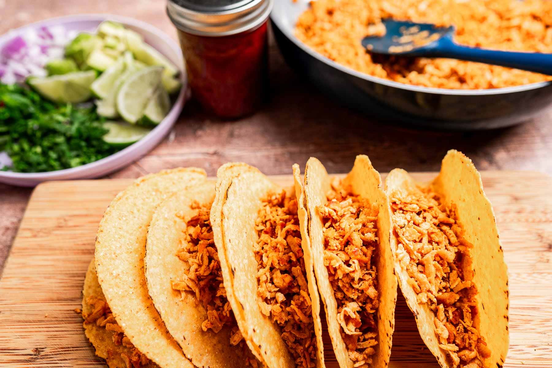 Tofu tacos filled with shredded chicken sit on a wooden board, accompanied by a plate of lime, cilantro, and onion. A skillet brimming with more savory filling beckons in the background.