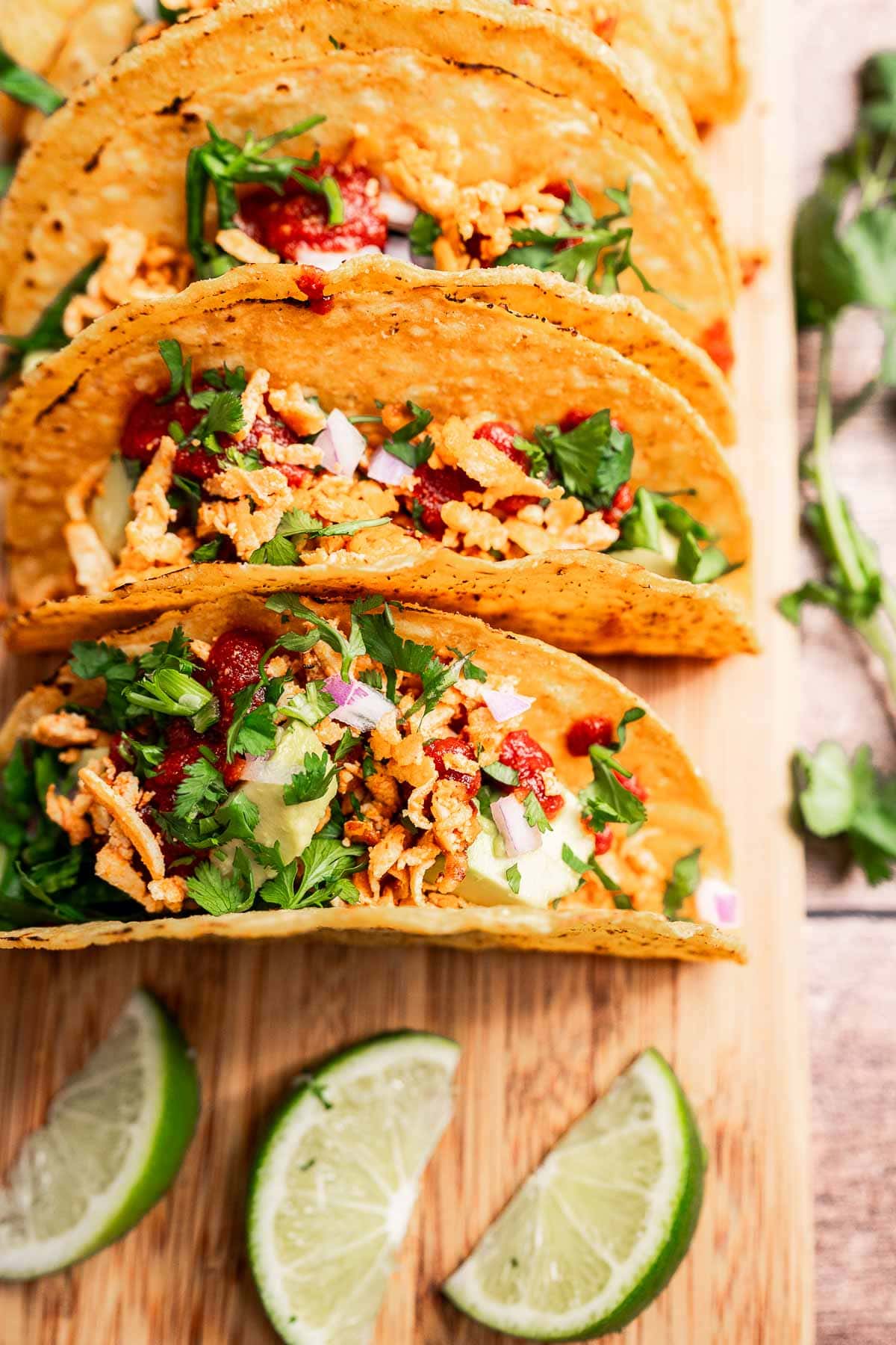 Three crispy tofu tacos filled with lettuce, diced tomatoes, onions, and shredded cheese on a wooden board. Lime wedges are placed in front.