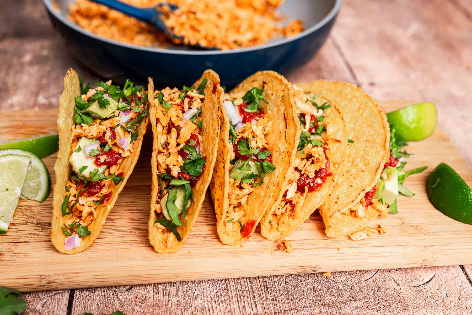 Four hard shell tacos filled with seasoned shredded chicken, diced avocado, chopped red onions, cilantro, and drizzled sauce sit enticingly on a wooden board alongside lime wedges. For a twist, try our tofu tacos for a delicious alternative.