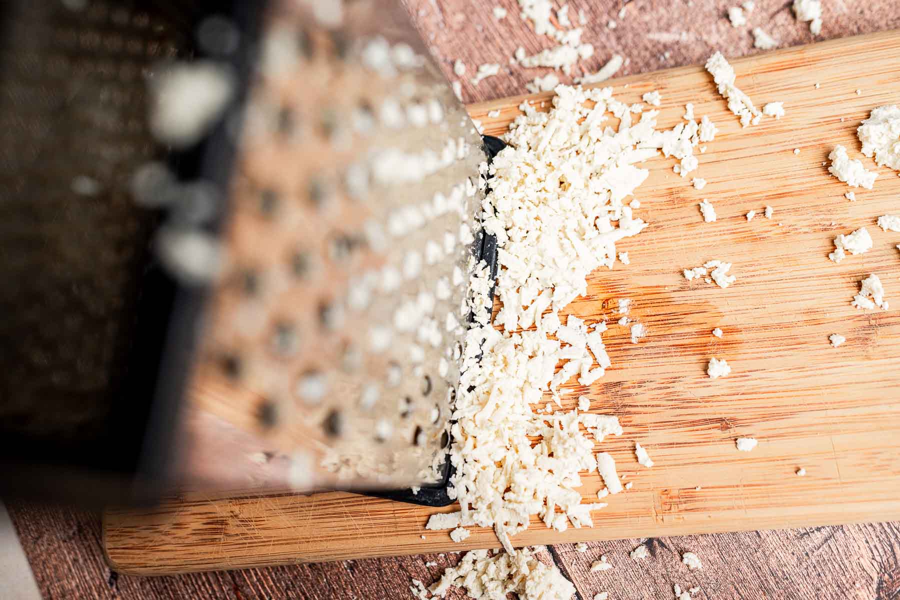 A metal box grater on its side with shredded cheese scattered on a wooden cutting board awaits the assembly of delicious tofu tacos.