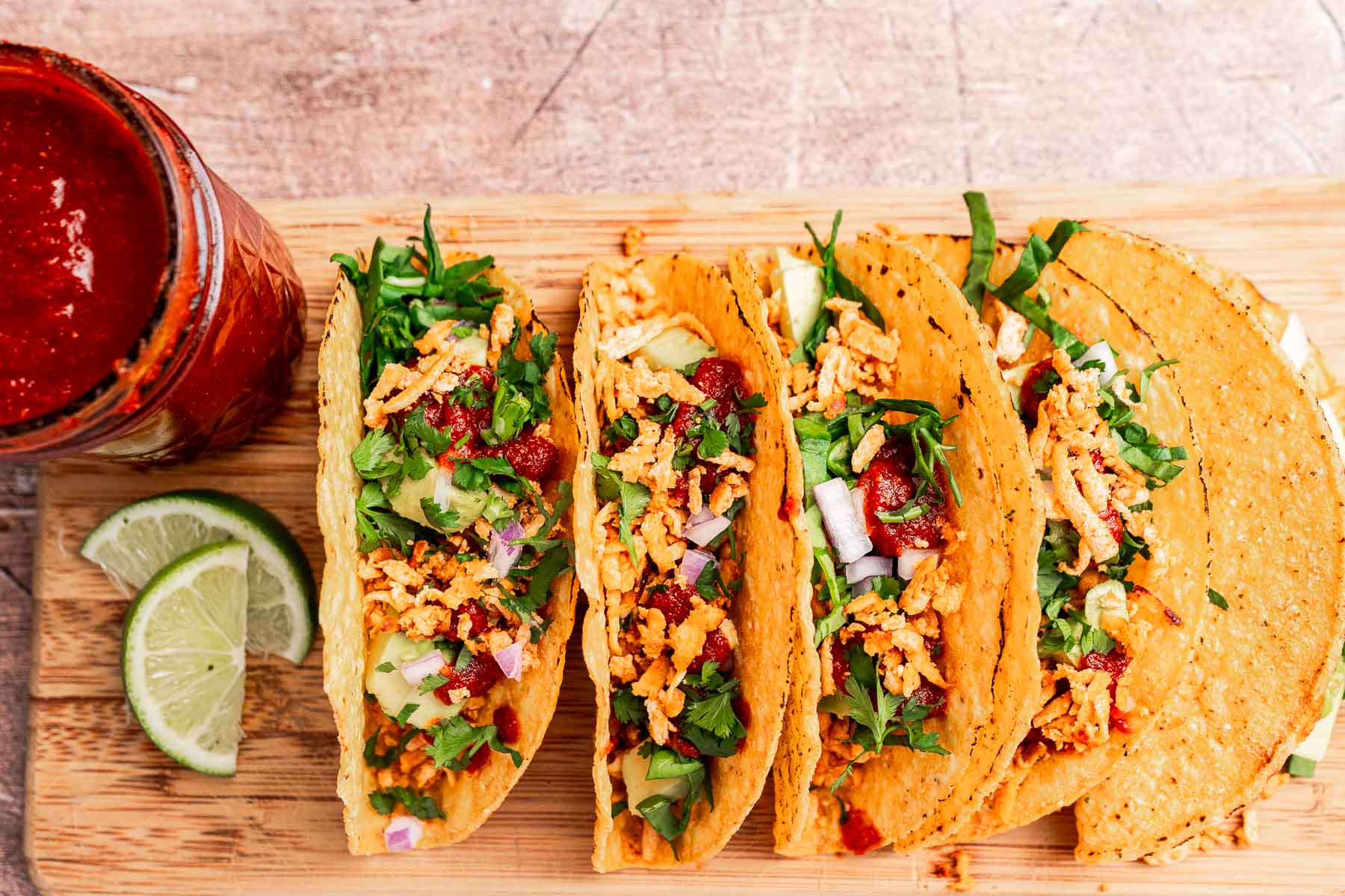 Four crispy tofu tacos filled with shredded meat, lettuce, onion, and salsa rest on a wooden board next to lime wedges and a jar of red sauce.