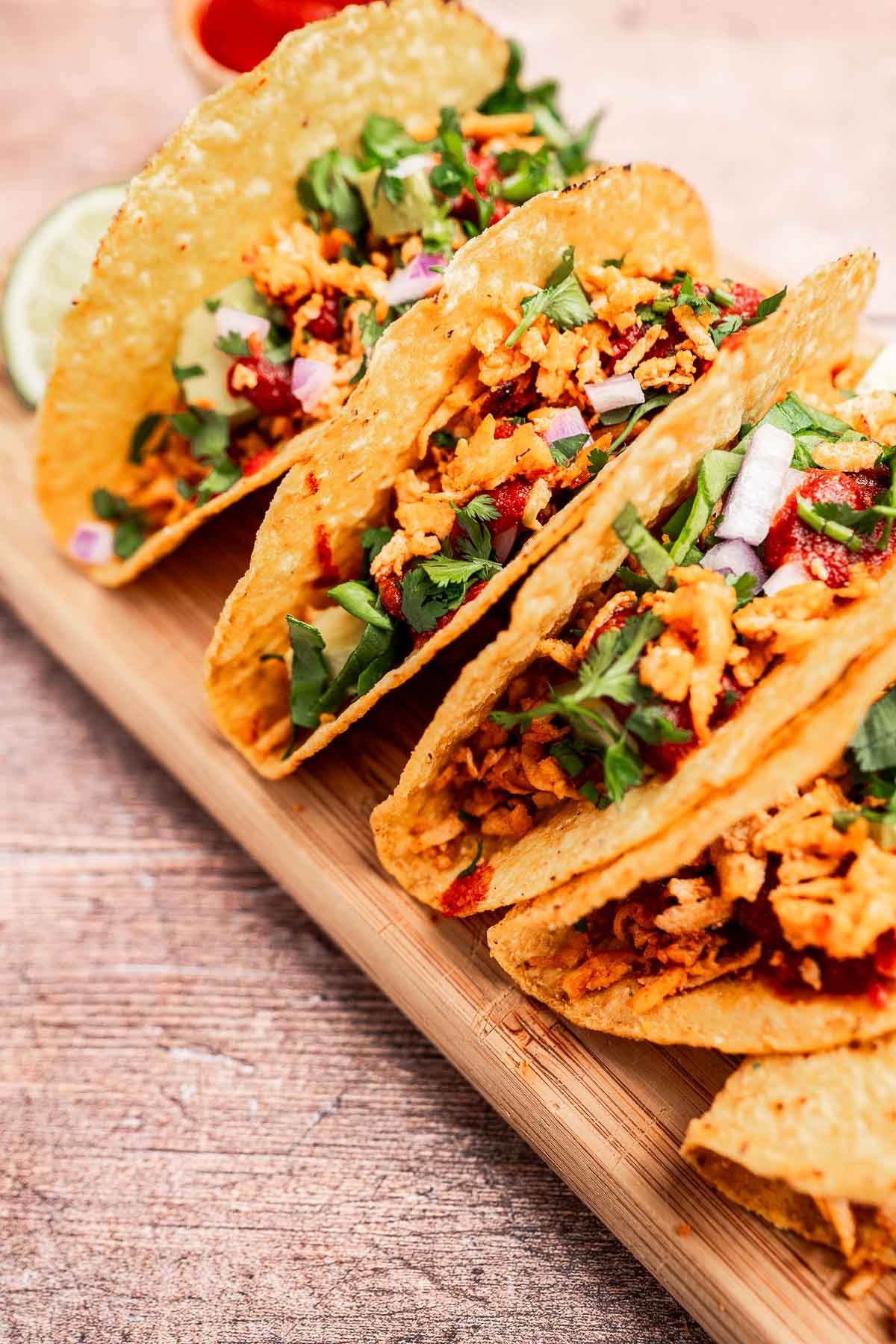 Three crispy tofu tacos filled with shredded meat, diced onions, cilantro, and sauce are artfully arranged on a wooden board.
