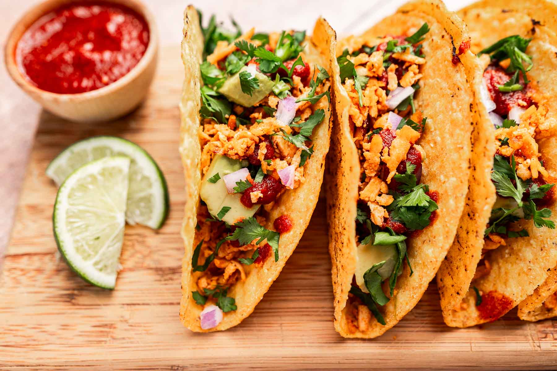 Three hard shell tofu tacos filled with shredded meat, diced onions, cilantro, and sauce, served on a wooden board with lime wedges and a bowl of red sauce.
