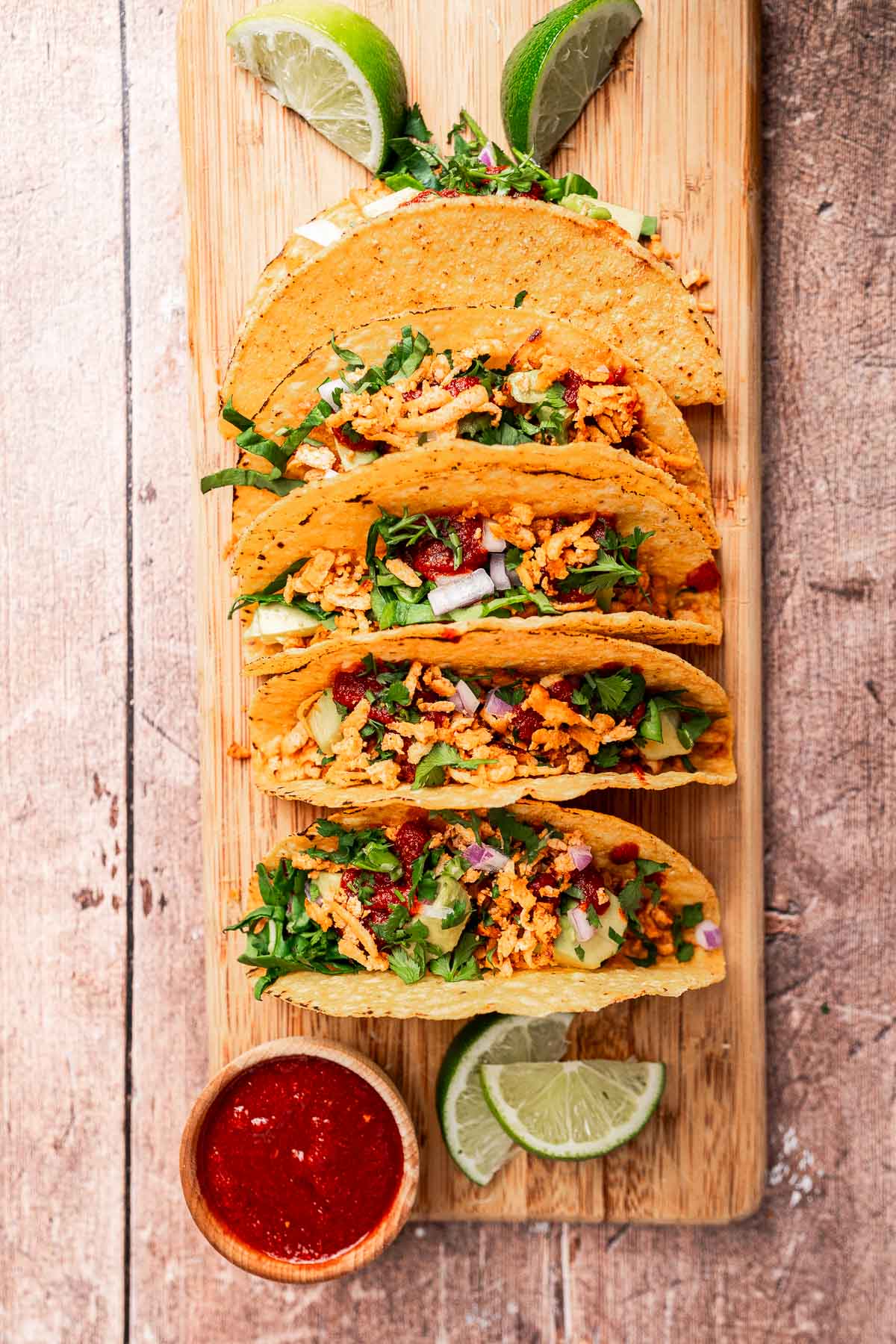 Four crispy tofu tacos filled with vegetables on a wooden board, garnished with lime wedges and accompanied by a small bowl of red sauce.