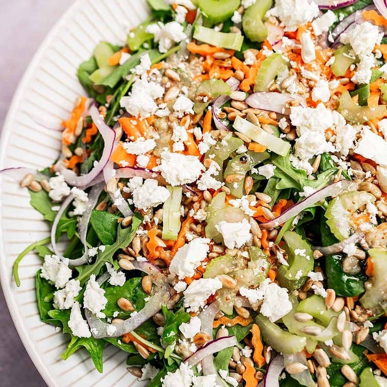A close-up of a fresh spinach and arugula salad with shredded carrots, cucumber slices, red onion, sunflower seeds, and crumbled feta cheese on a white plate.