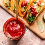 A jar of red salsa and a spoon rest beside three tacos on a wooden board, filled with greens, onions, and taco sauce. Lime wedges are also scattered across the board.