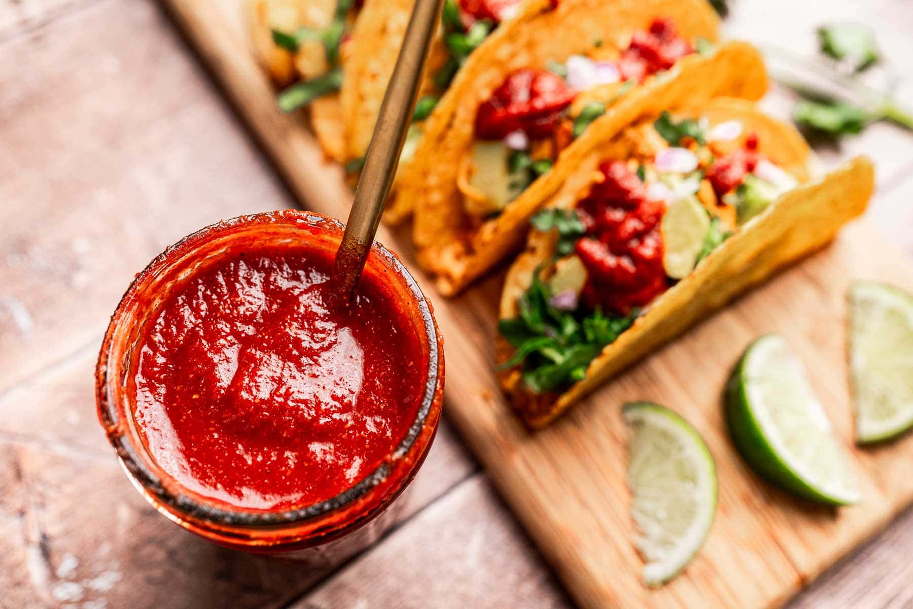 A jar of red salsa and taco sauce sits invitingly beside three tacos on a wooden board, garnished with fresh onions and herbs. Lime wedges add a zesty touch to the vibrant presentation.