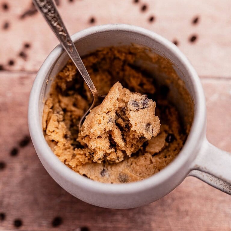 A mug filled with chocolate chip cookie dough and a spoon inside, perched on a wooden surface sprinkled with scattered chocolate chips, offers the allure of an oat flour mug cake waiting to be devoured.