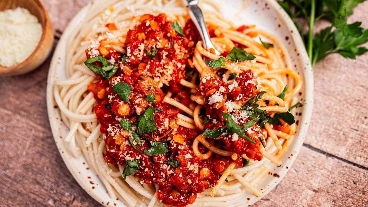 A plate of spaghetti topped with tomato sauce, chopped parsley, and grated cheese, with a fork twirling some noodles. A parsley bunch and a small bowl of cheese are nearby.
