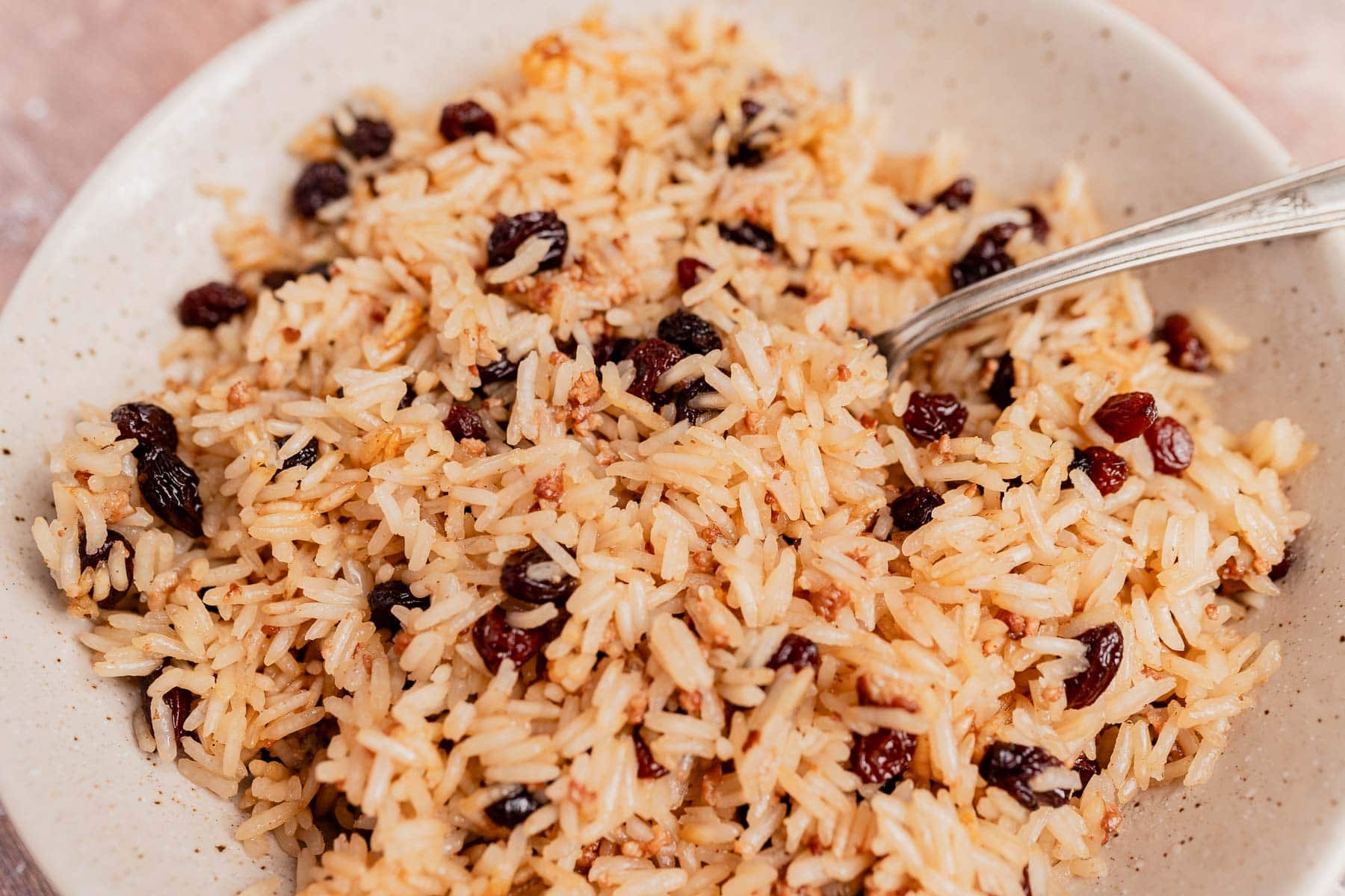 A close-up of a bowl of Arroz con Coco—cooked rice mixed with raisins—captured in detail, with a metal spoon partially visible on the right side.