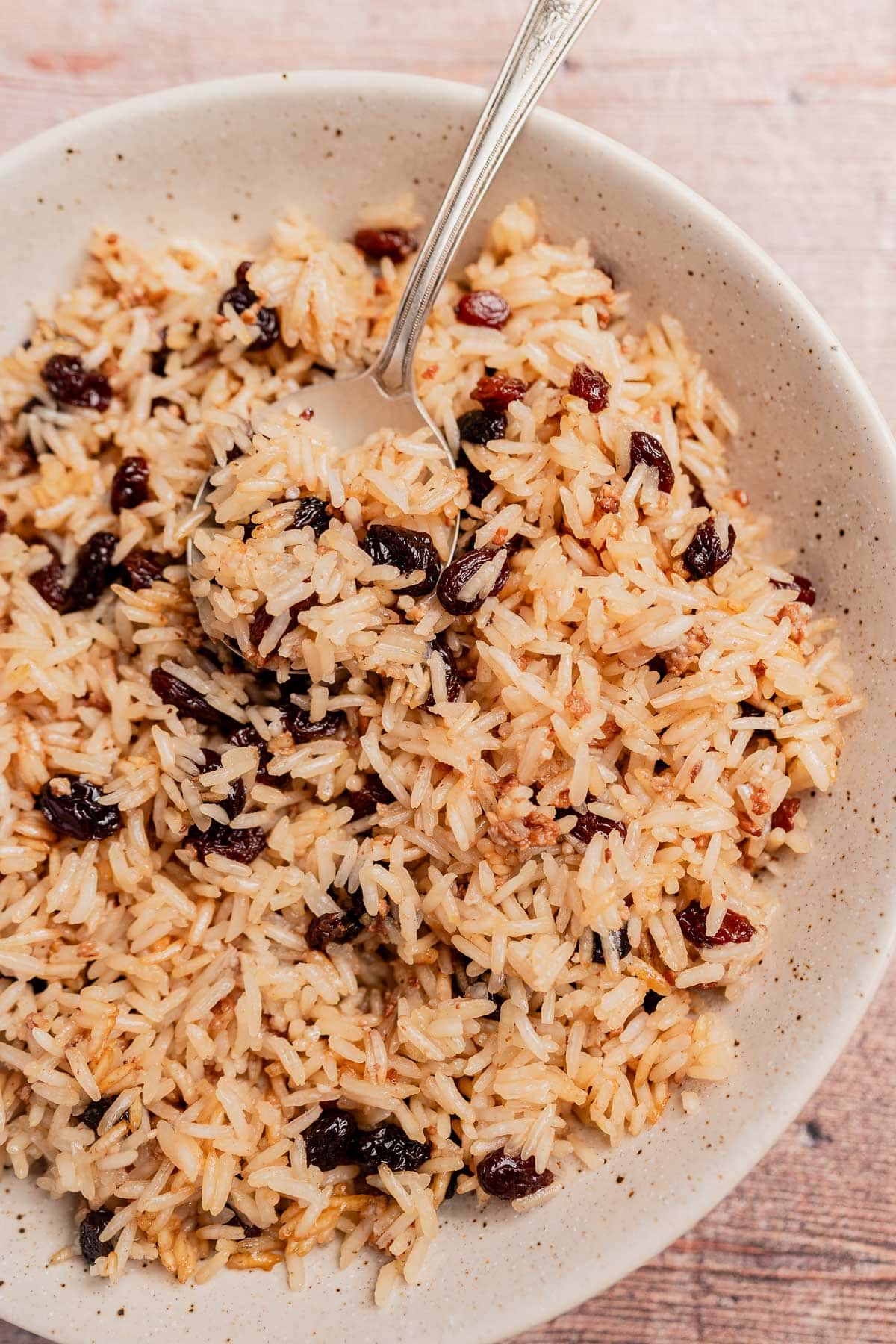 A bowl of Arroz con Coco, cooked rice mixed with raisins, sits on a wooden surface with a spoon resting inside.