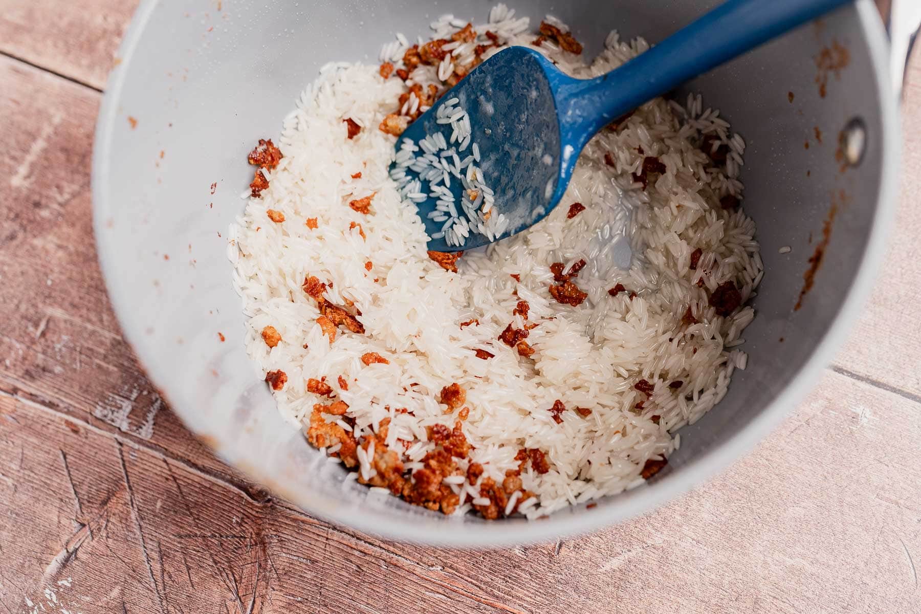 A pot containing uncooked white rice and browned bits, often seen in dishes like Arroz con Coco, being stirred with a blue spatula on a wooden surface.