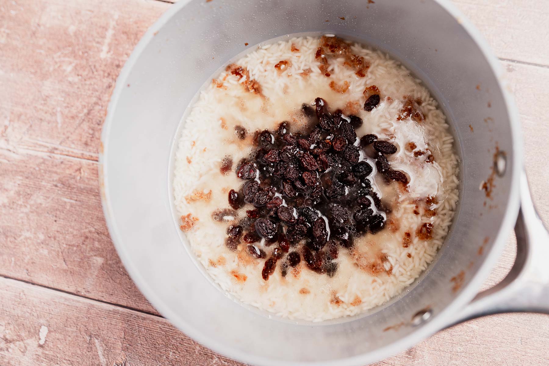 Arroz con Coco ingredients—uncooked rice, raisins, and liquid—sit together in a saucepan on a wooden surface, ready to be transformed into this classic dish.