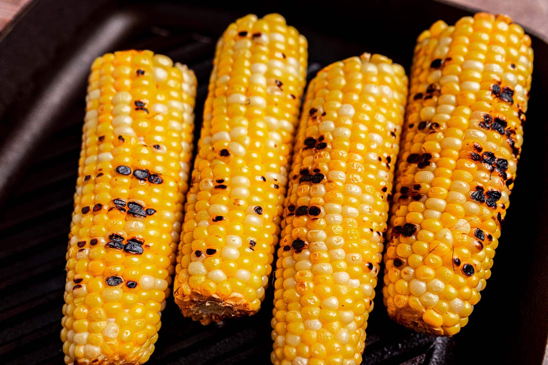 Four ears of grilled corn with visible char marks lined up on a grill pan, perfect for making a delicious charred corn salad.