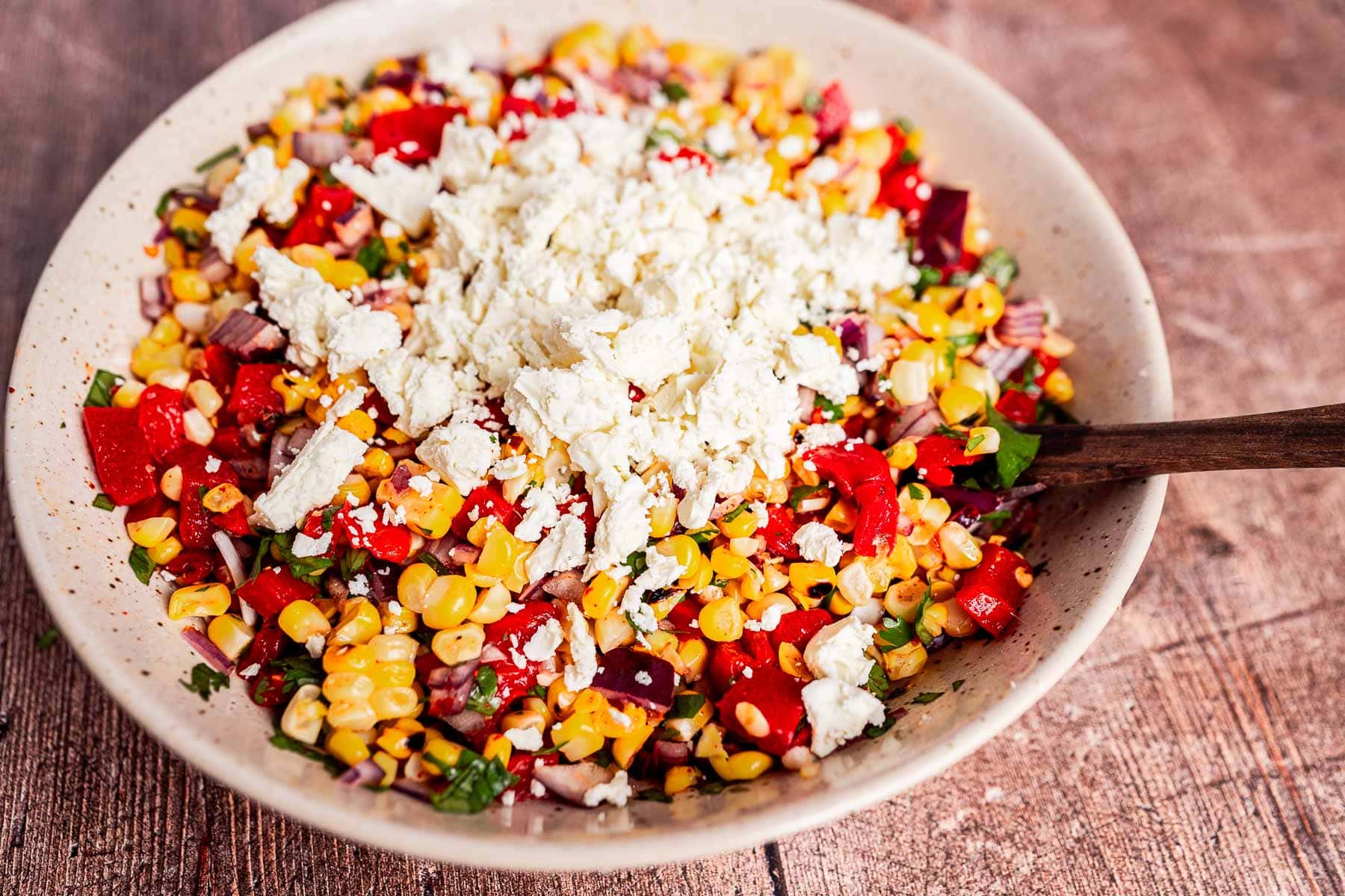 A bowl of charred corn salad with chopped tomatoes, red onion, herbs, and a generous topping of crumbled feta cheese, served with a wooden spoon.