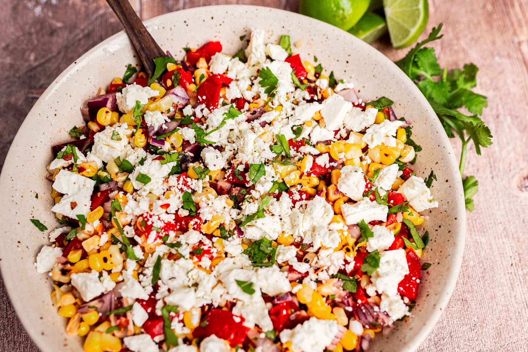 A bowl of charred corn salad with chopped red peppers, onions, cilantro, and crumbled white cheese, served with lime wedges and extra cilantro on the side.