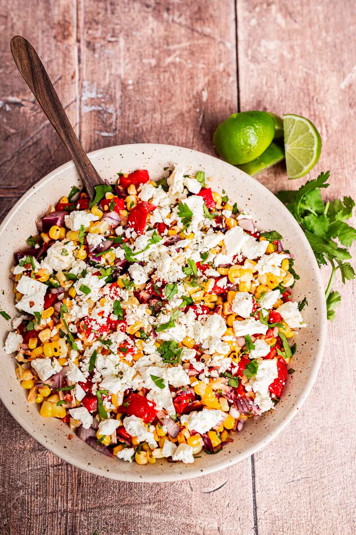 A bowl of charred corn salad with feta cheese, chopped red onion, tomatoes, cilantro, and a wooden spoon sits next to a halved lime and fresh cilantro on a wooden surface.