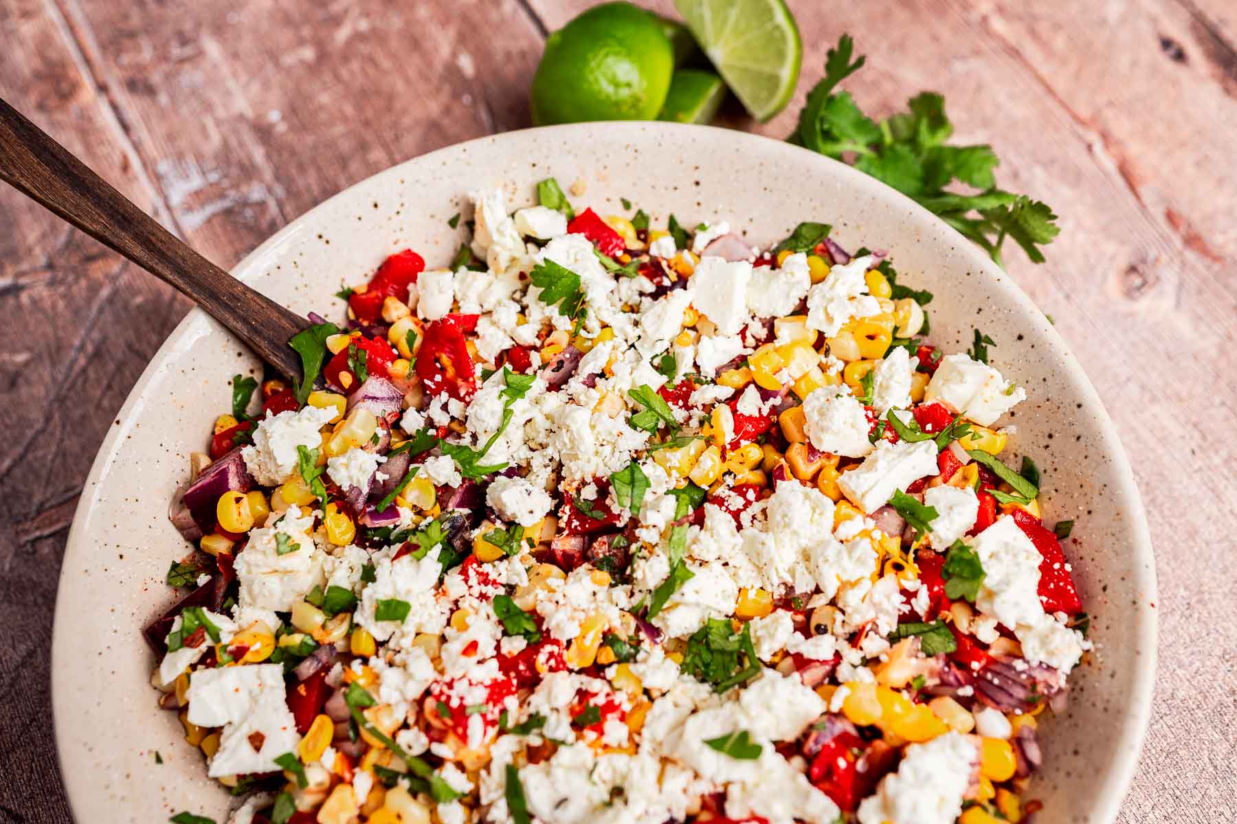 A bowl of charred corn salad with tomatoes, red onions, chopped herbs, and crumbled feta cheese, accompanied by a wooden spoon, cilantro, and lime halves on a rustic wooden surface.