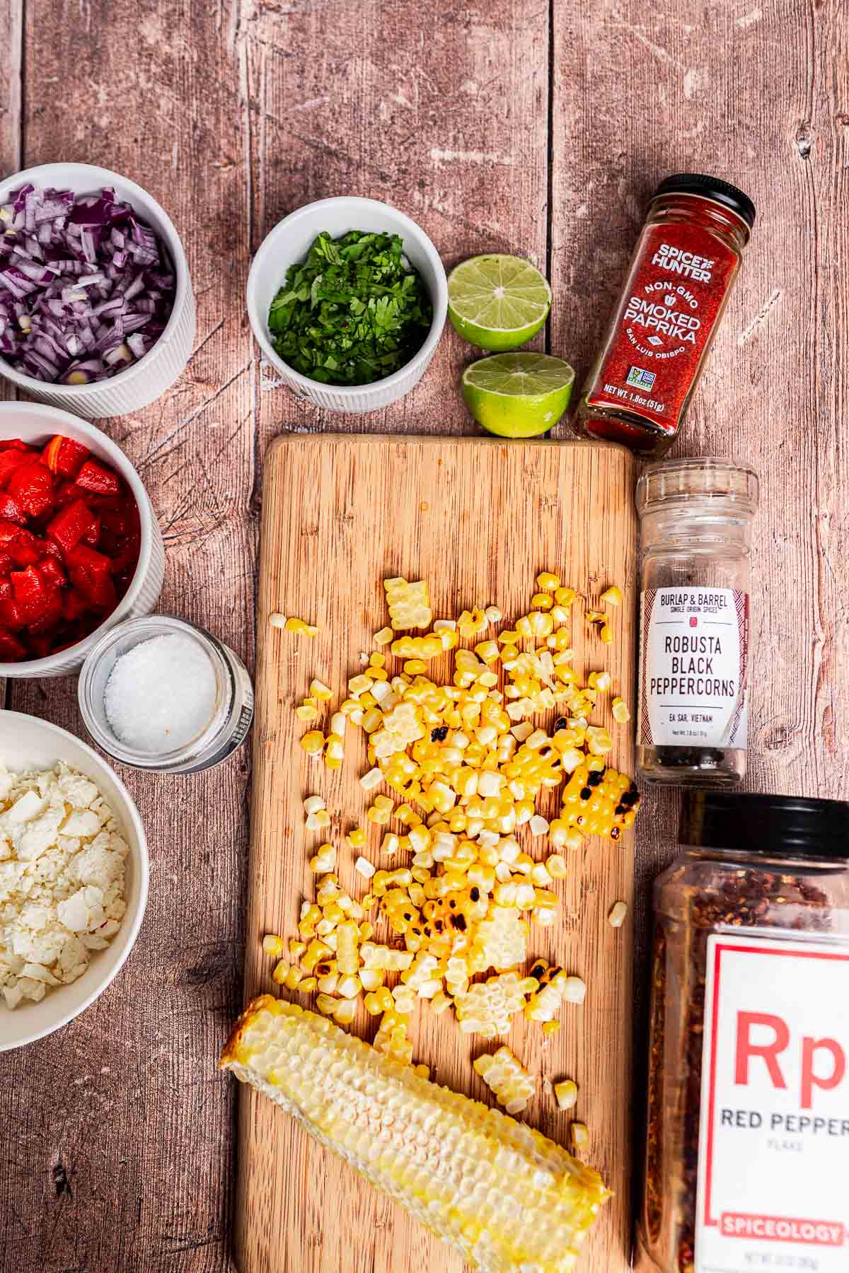 Chopped grilled corn for charred corn salad on a cutting board, surrounded by bowls of feta, red onion, red pepper, cilantro, spices, salt, and a halved lime on a wooden surface.