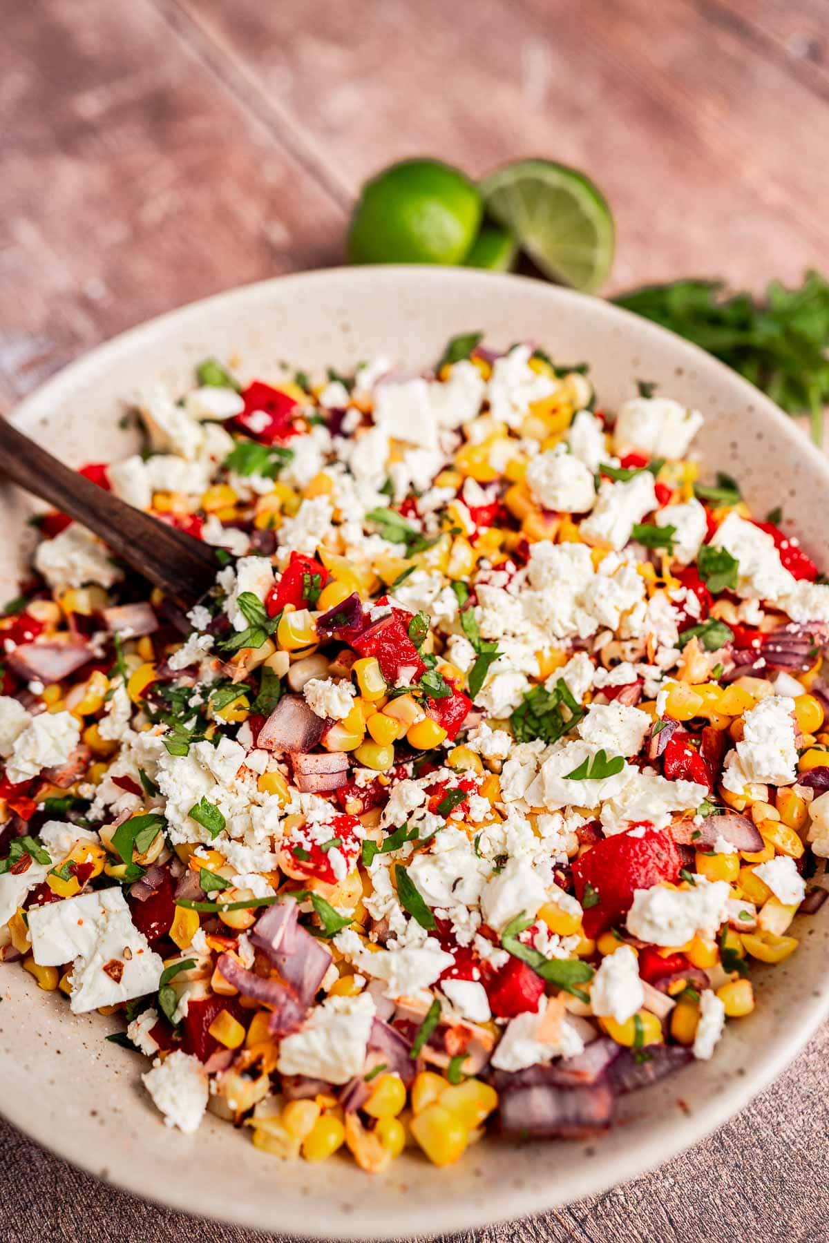 A bowl of charred corn salad with chopped red peppers, onions, cilantro, and crumbled white cheese, with a serving spoon and limes in the background.