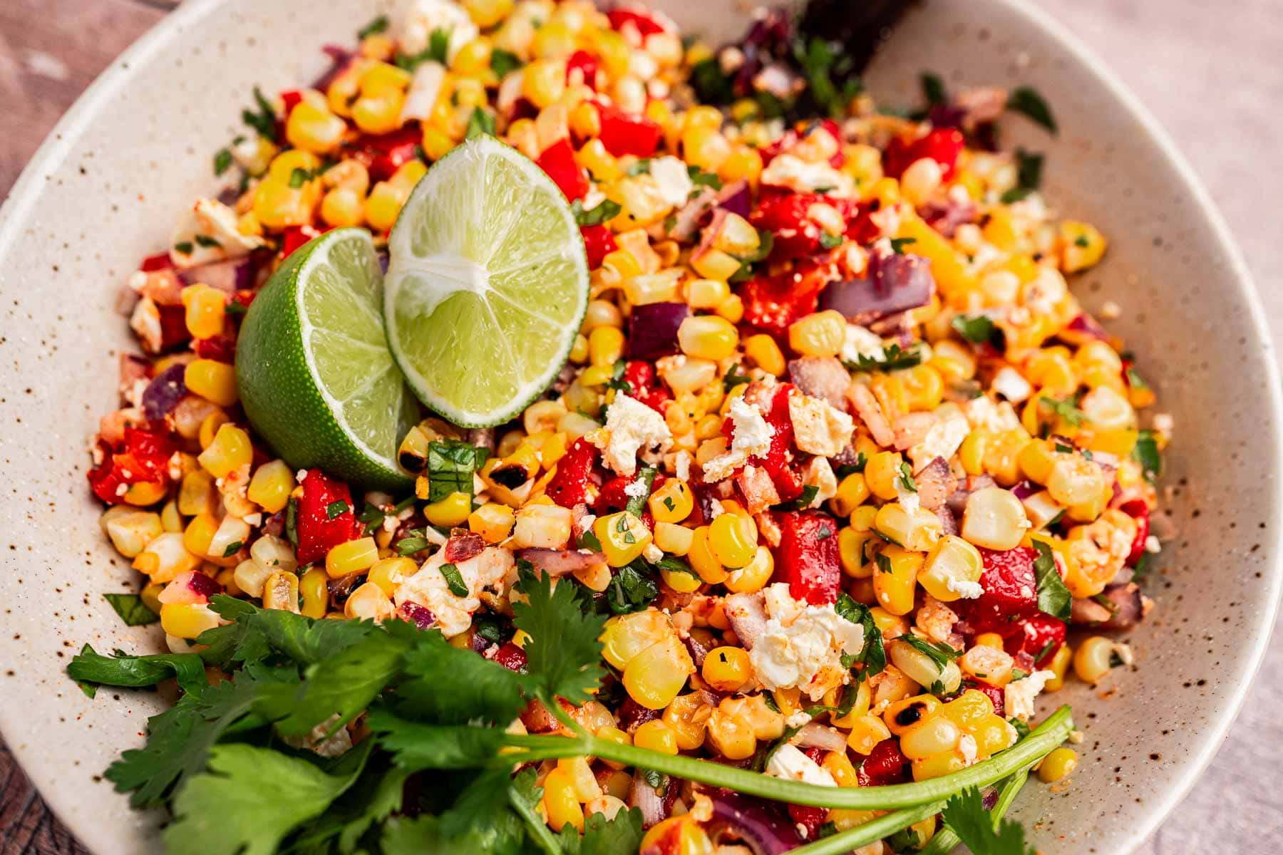 A bowl of charred corn salad with red peppers, crumbled cheese, cilantro, and lime wedges brings the flavors of Mexican street corn to your table.