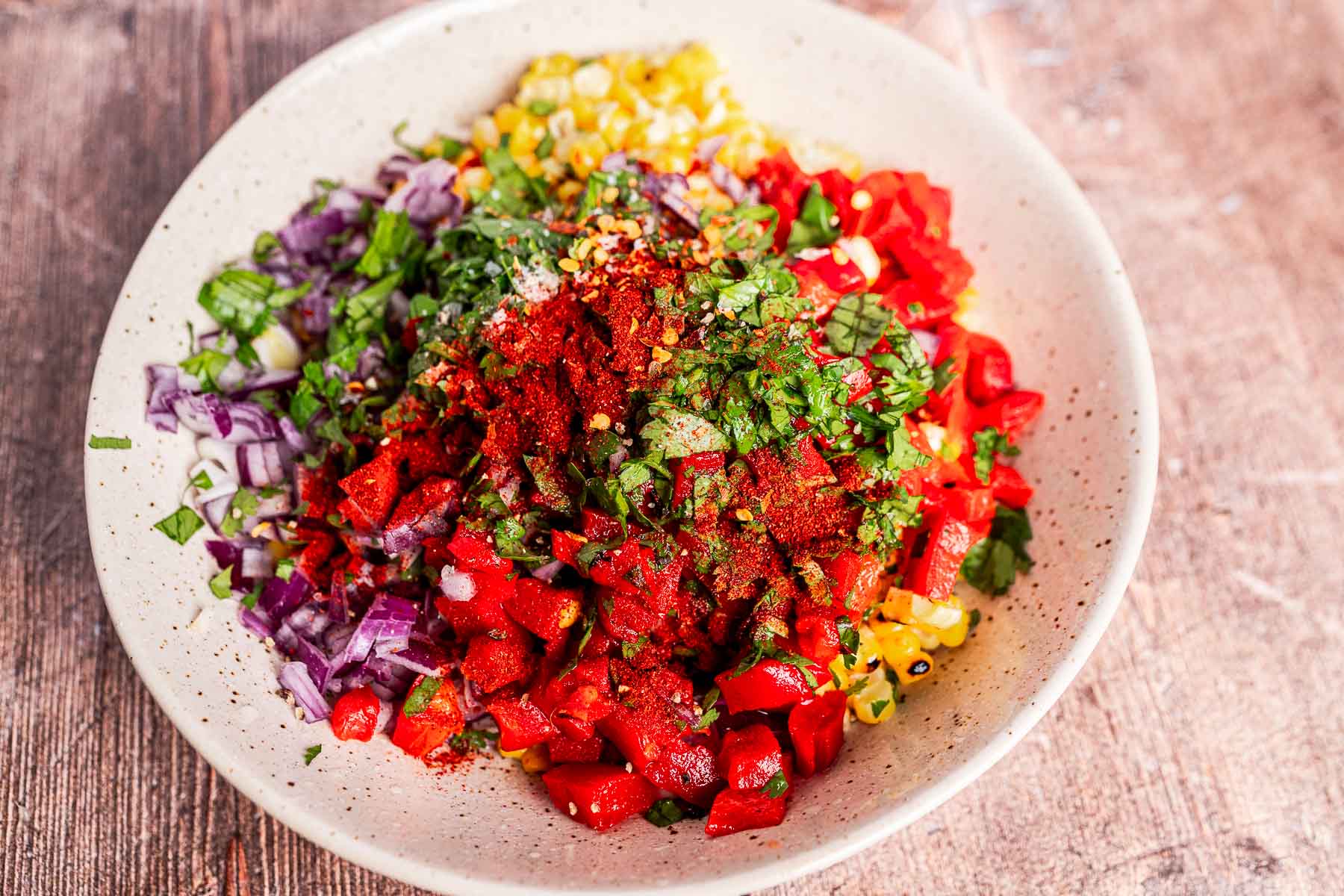 A bowl filled with chopped red onion, charred corn, tomatoes, herbs, and spices, ready to be mixed for a fresh salsa or charred corn salad.