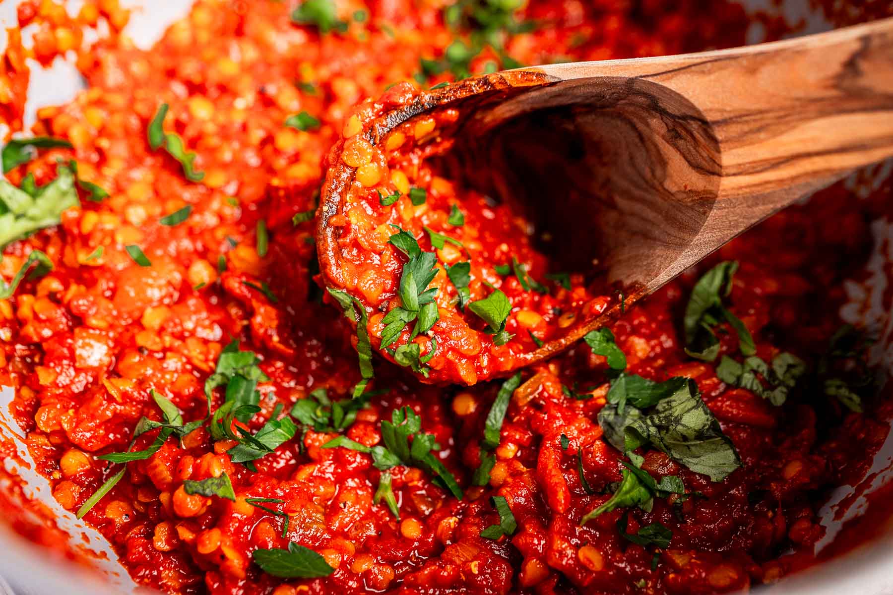A wooden spoon stirs a chunky red vegetarian bolognese sauce with lentils and chopped fresh herbs.