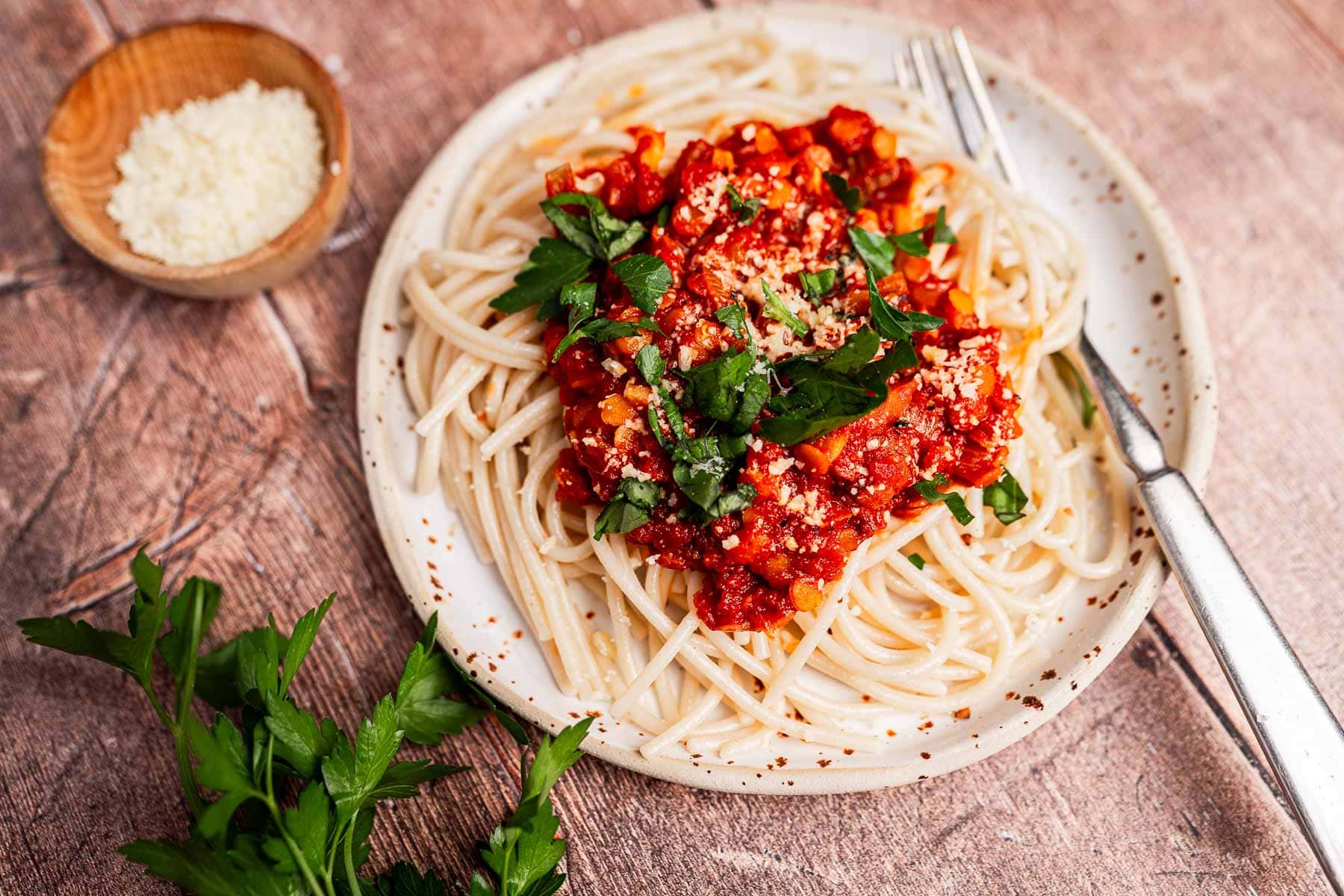 A plate of vegetarian bolognese-topped spaghetti with tomato sauce and fresh parsley sits next to a fork, knife, grated cheese in a small bowl, and a sprig of parsley on a wooden surface.
