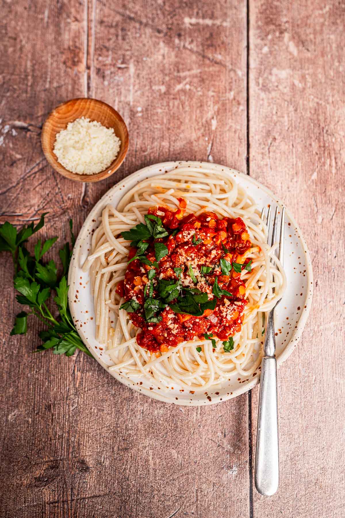 A plate of vegetarian bolognese spaghetti topped with tomato sauce and chopped parsley, served with a fork and a small bowl of grated cheese on a wooden table.