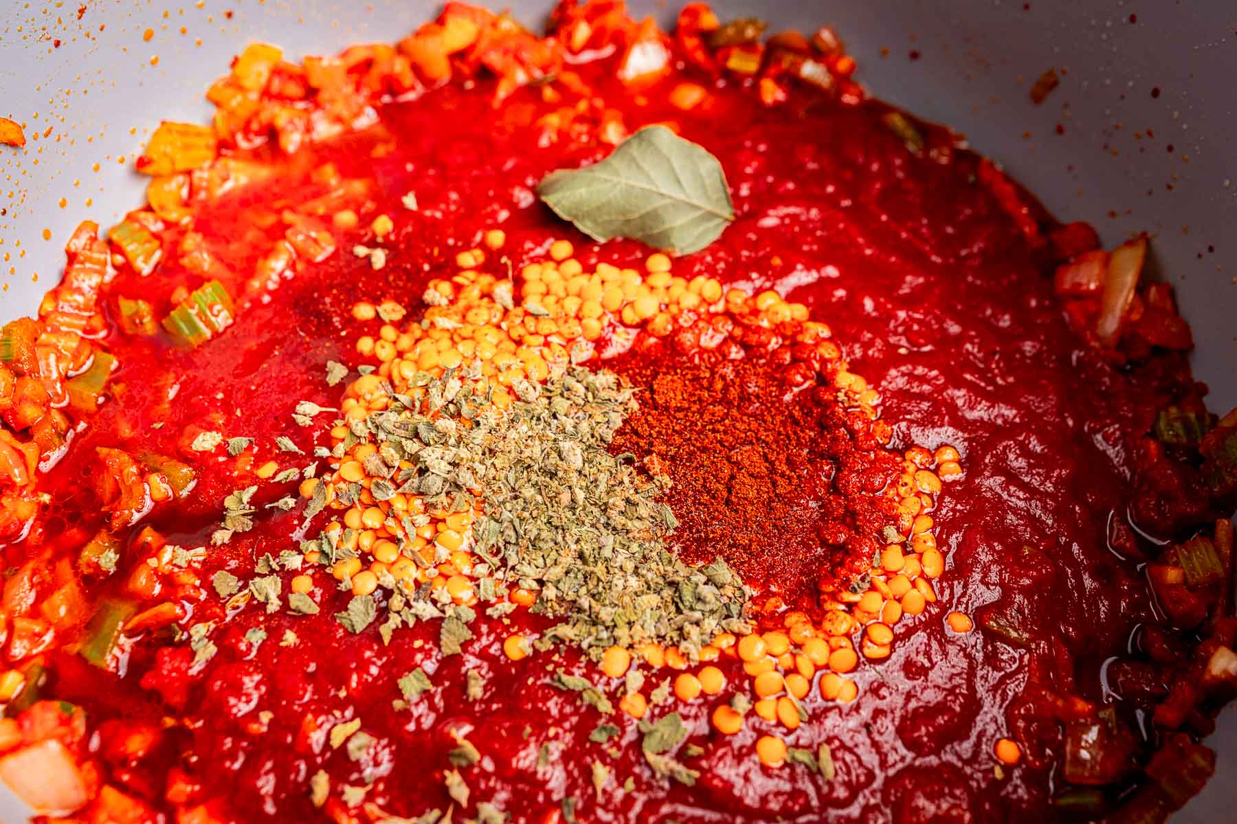 A close-up of vegetarian bolognese sauce in a pot with red lentils, dried herbs, paprika, and a bay leaf, surrounded by chopped celery and onions.