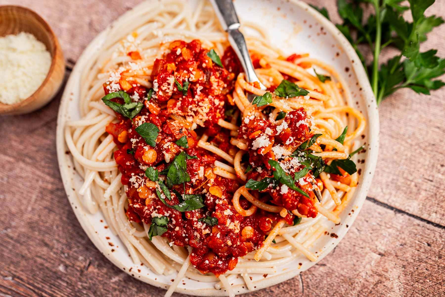 A plate of spaghetti topped with vegetarian bolognese, grated cheese, and chopped parsley, with a fork twirling some pasta. A bowl of parmesan and fresh parsley are nearby.