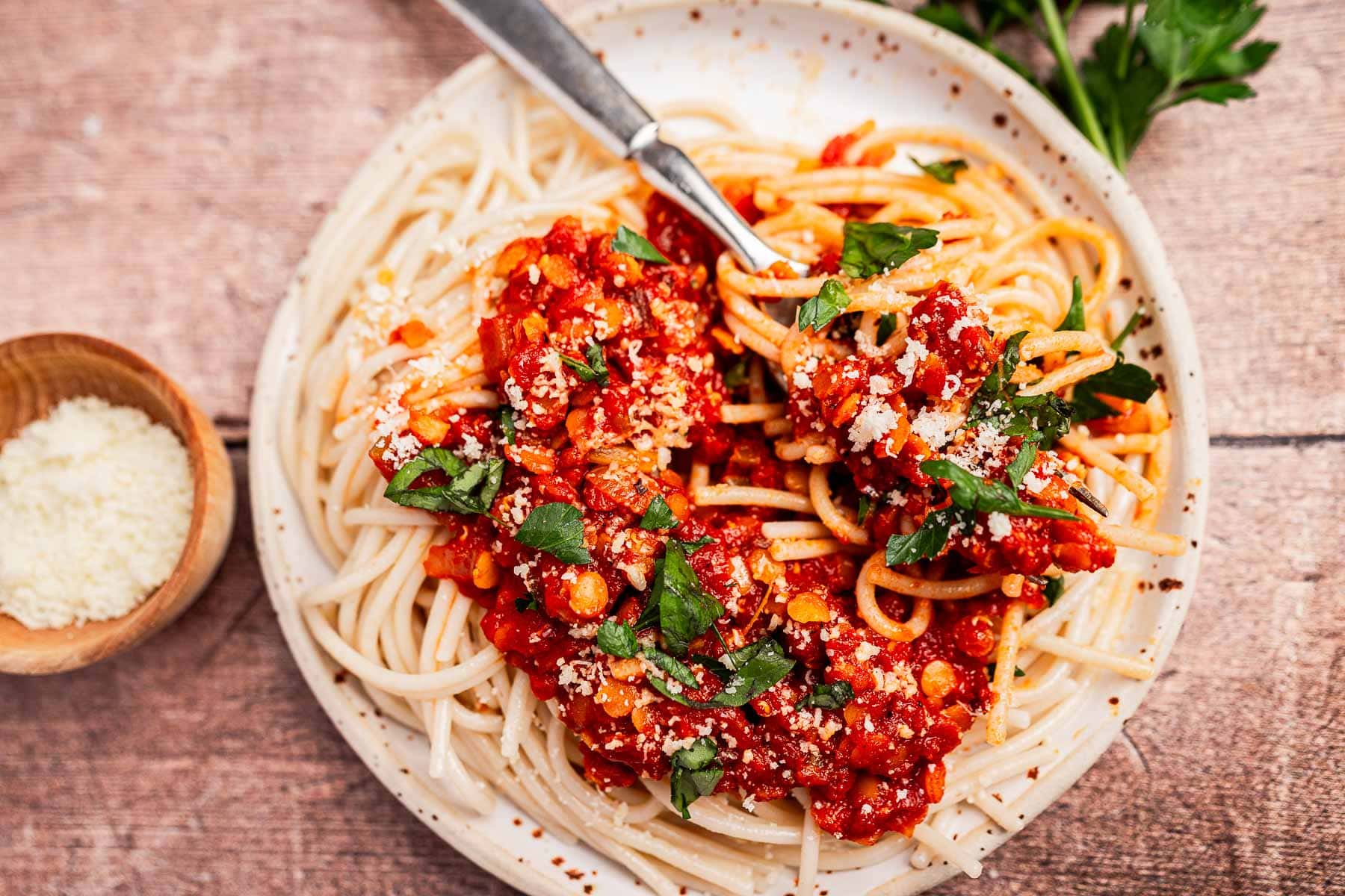 A plate of spaghetti topped with hearty vegetarian bolognese sauce, chopped parsley, and grated cheese, with a fork on the side. A small bowl of grated cheese is next to the plate.