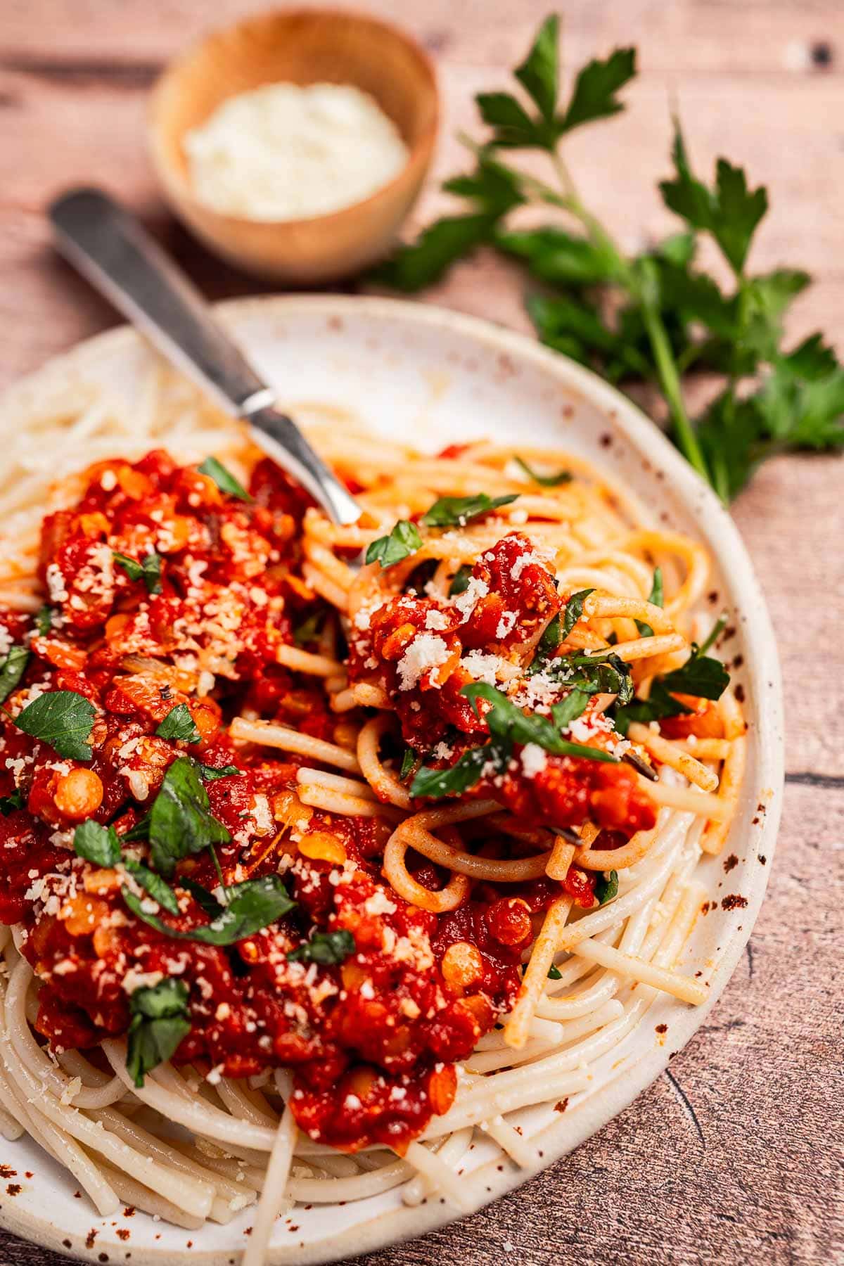 A plate of spaghetti topped with vegetarian bolognese sauce, chopped parsley, and grated cheese, with a fork and a bowl of extra cheese in the background.