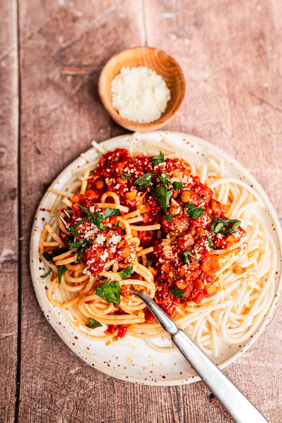 A plate of spaghetti topped with rich vegetarian bolognese, grated cheese, and fresh herbs, with a fork on the plate and a small wooden bowl of grated cheese in the background.