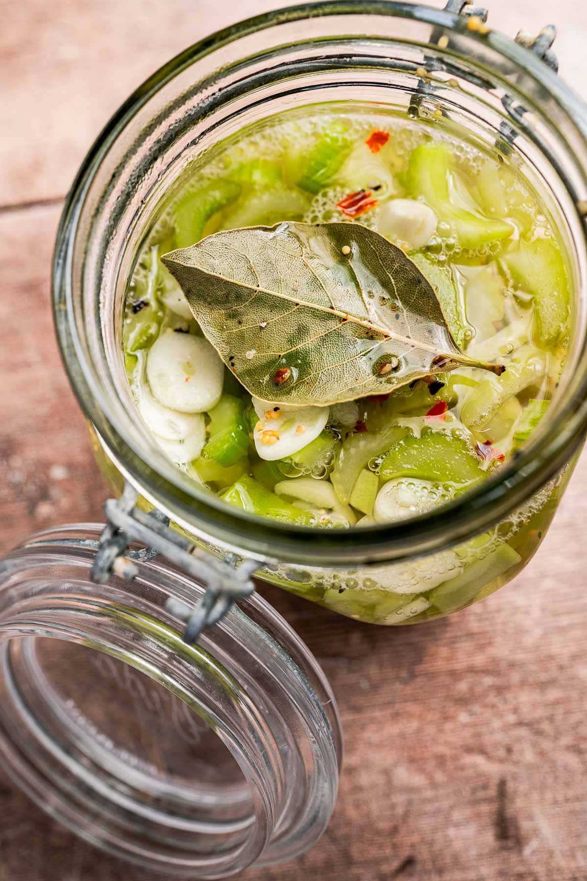 An open glass jar filled with pickled vegetables, including pickled celery, sliced green peppers, garlic, spices, and topped with a bay leaf.