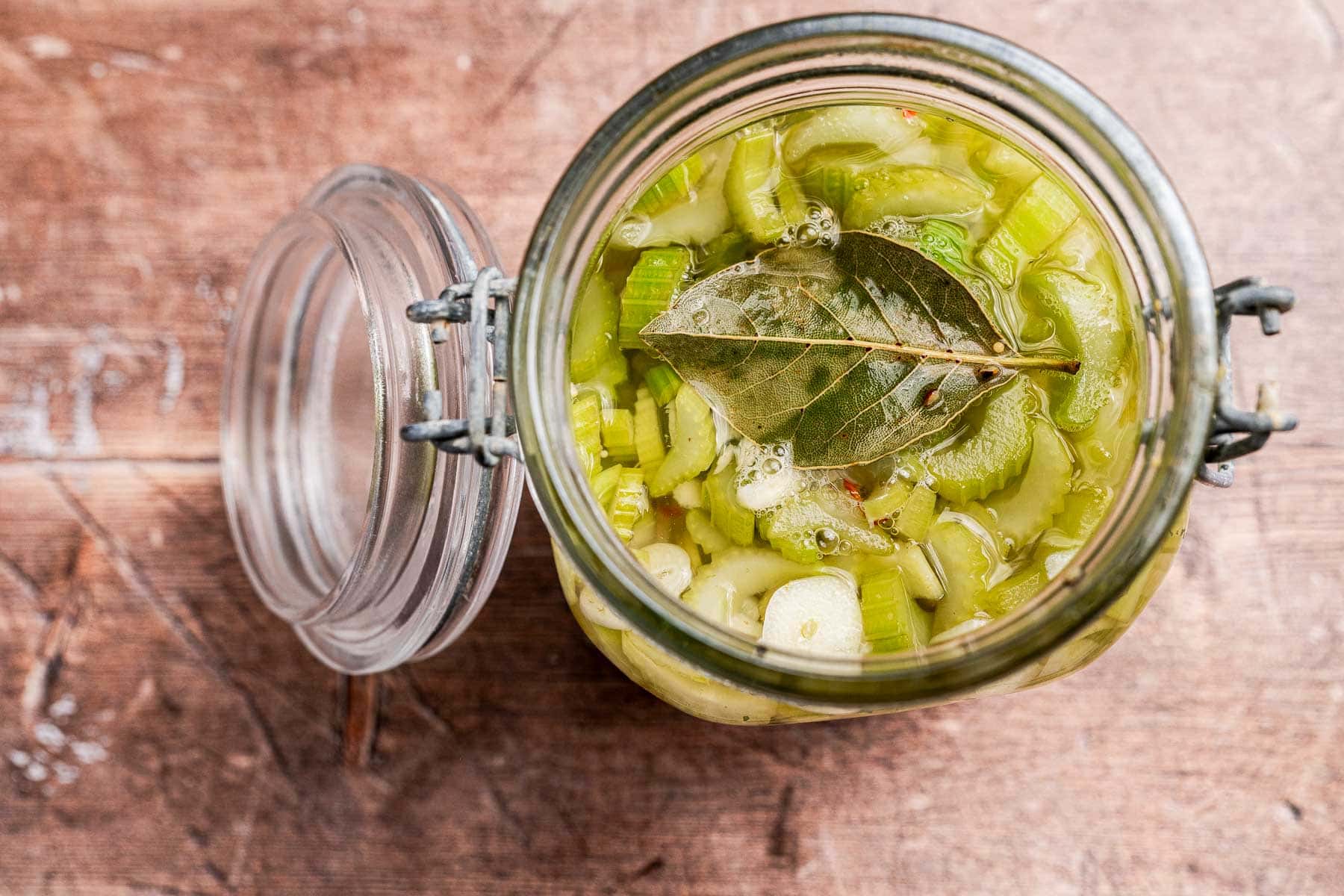 An open glass jar filled with pickled celery, sliced garlic, spices, and a bay leaf sits on a wooden surface.