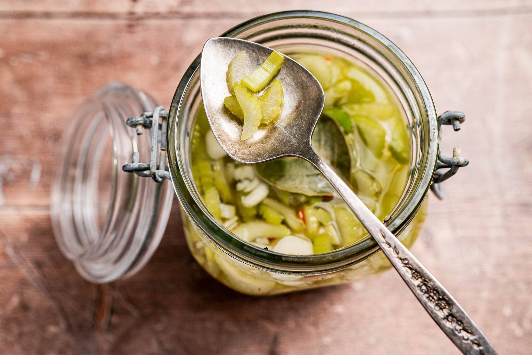 A spoon holding sliced pickled vegetables, including tangy pickled celery, hovers above an open glass jar brimming with more pickled vegetables on a wooden surface.