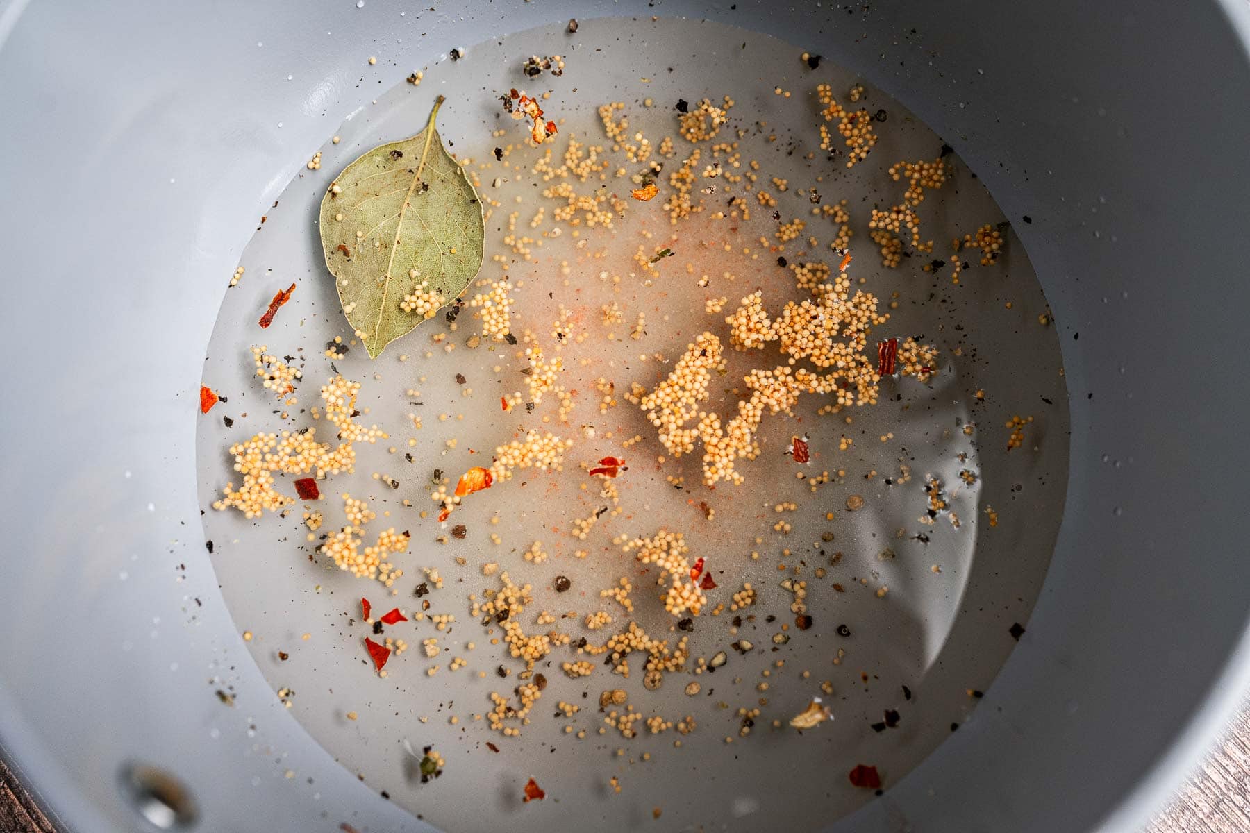 A close-up of a pot with water, a bay leaf, mustard seeds, peppercorns, and crushed red pepper flakes prepared for pickled celery.